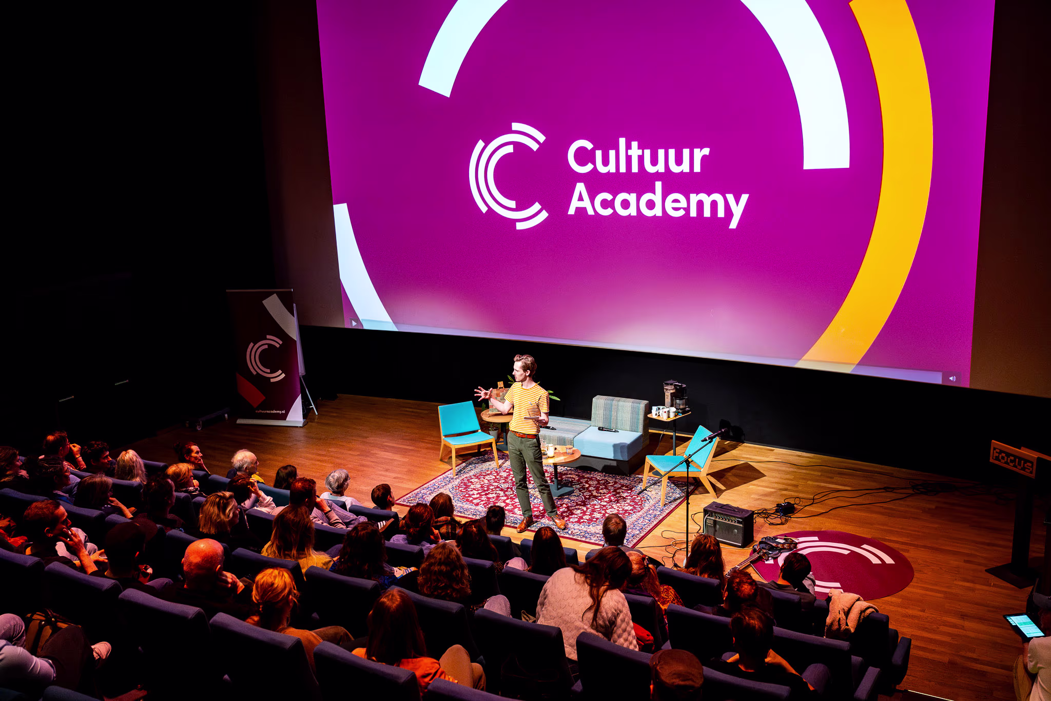 Een spreker presenteert op het podium voor een volle zaal, met het logo van Cultuur Academy op de grote scherm achter hem.