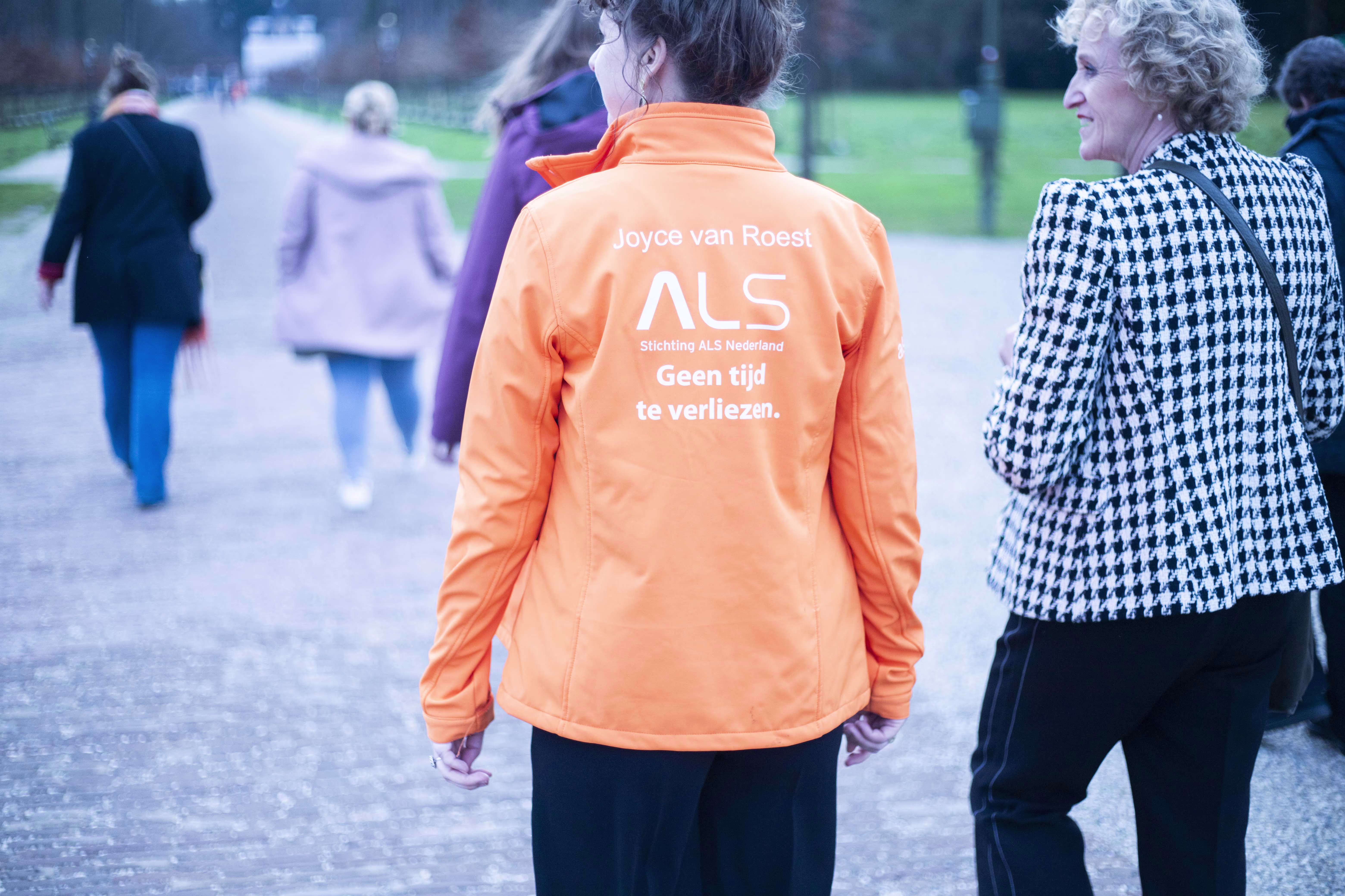 Persoon in oranje ALS Nederland jas loopt samen met anderen tijdens een wandelevenement voor het goede doel.