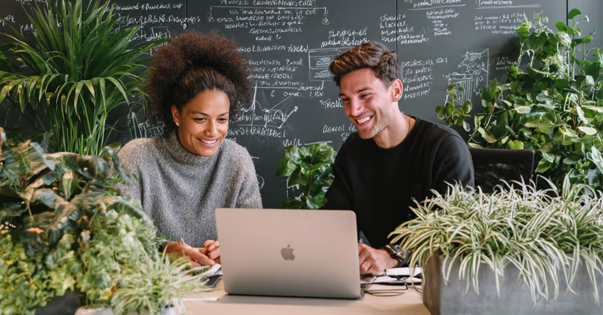Twee collega's werken lachend samen achter een laptop in een groen kantoor met planten en een schoolbord op de achtergrond.