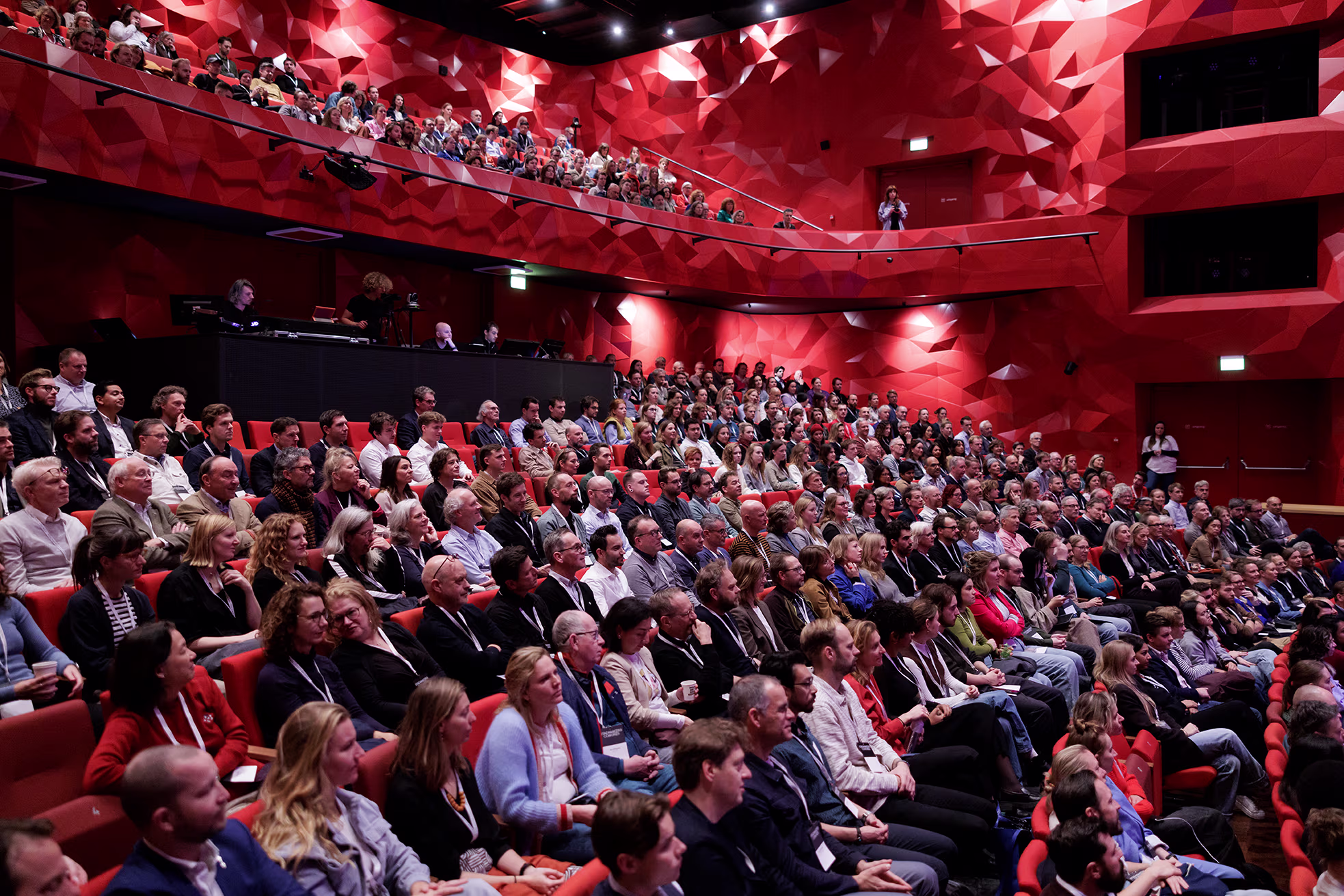 Een volle, moderne theaterzaal met rode wanden waar een grote groep mensen naar een presentatie kijkt tijdens een evenement.