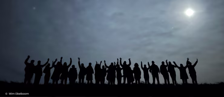 Silhouetten van een groep mensen met opgestoken armen tegen een bewolkte hemel met zon, uitend vreugde en saamhorigheid.
