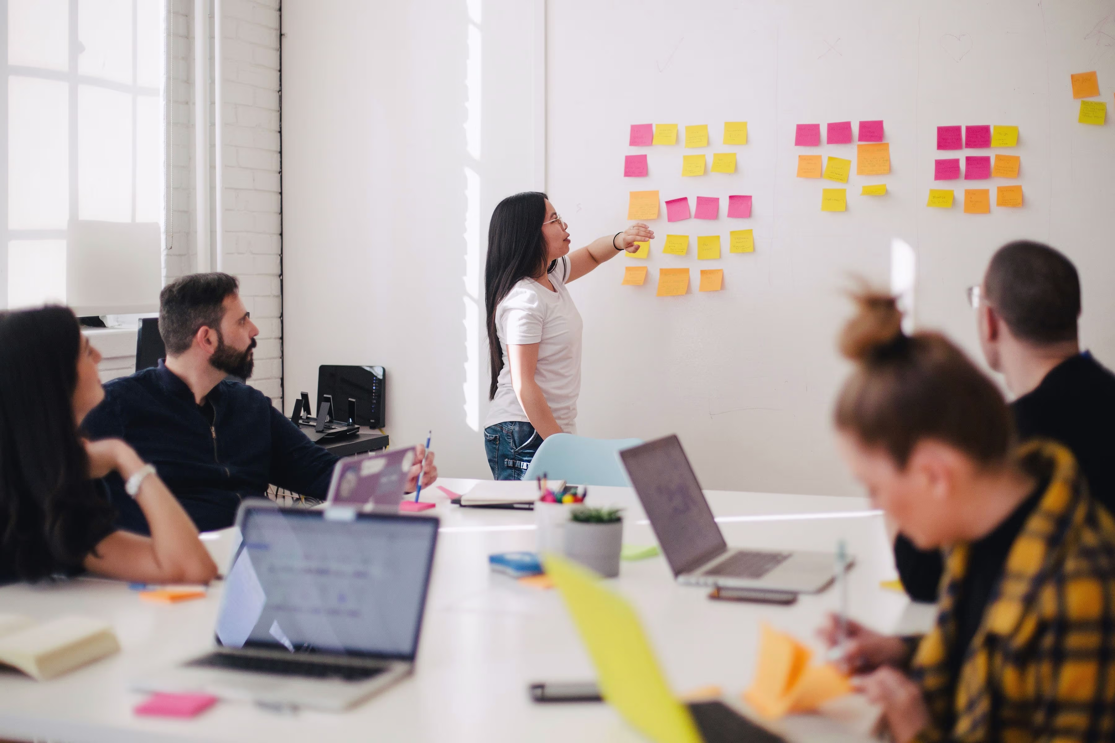 Een vrouw presenteert aan een whiteboard met gekleurde post-its terwijl collega's met laptops aan een tafel zitten te luisteren.