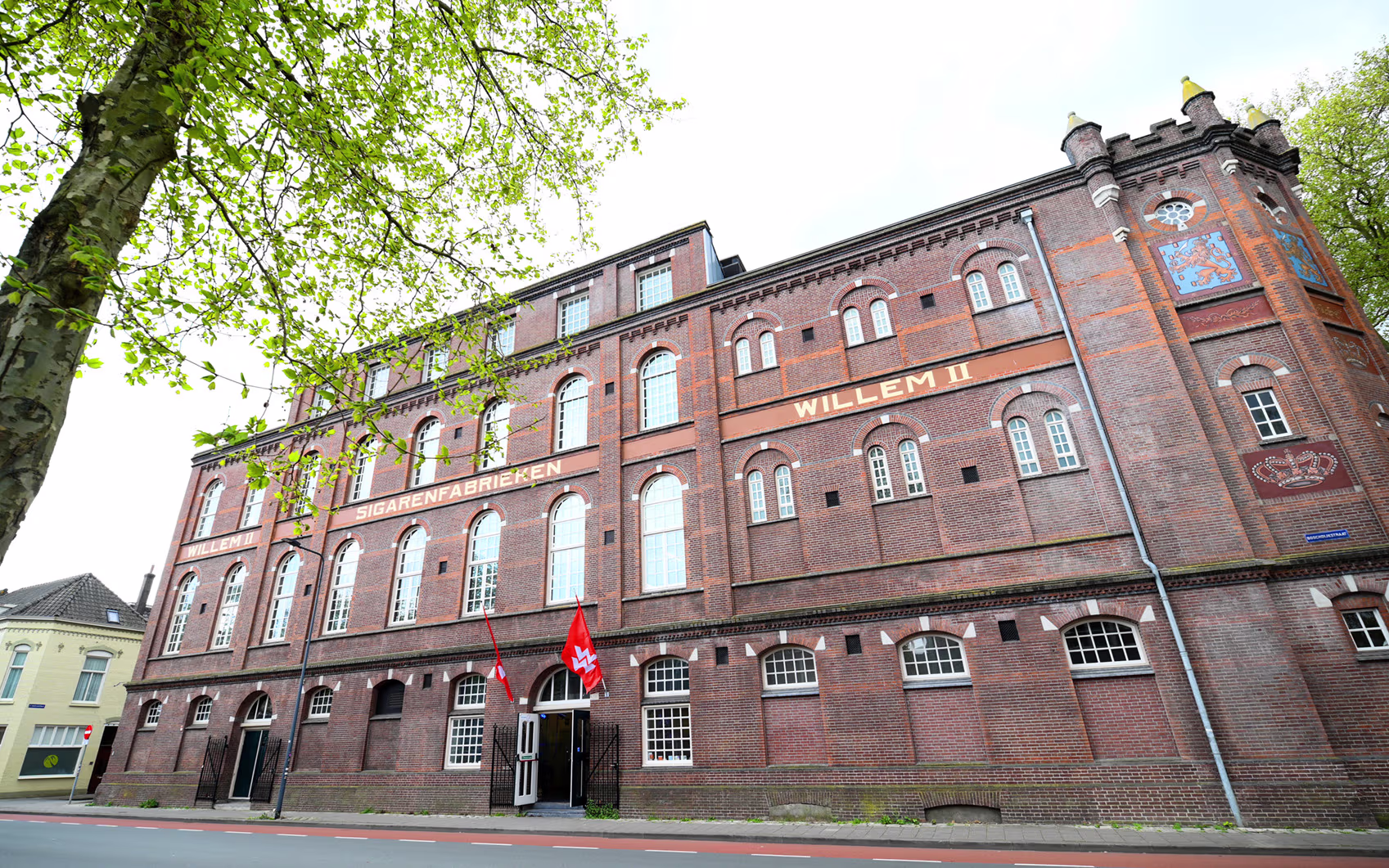 Een historisch rood bakstenen gebouw met grote ramen en de tekst "WILLEM II" op de gevel, omgeven door groene bomen.