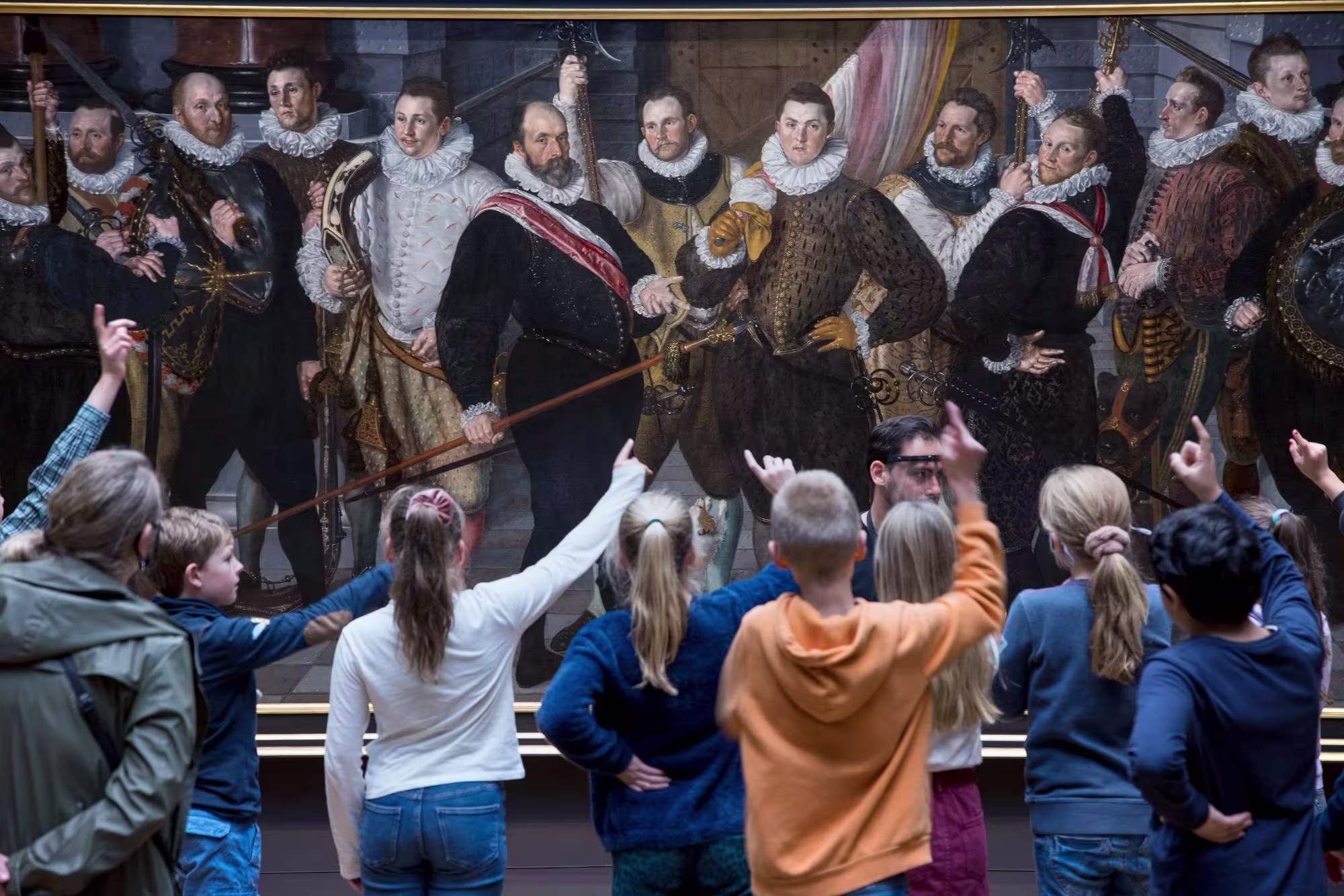 Kinderen wijzen enthousiast naar een historisch groepsportret in het Rijksmuseum tijdens een museumbezoek.