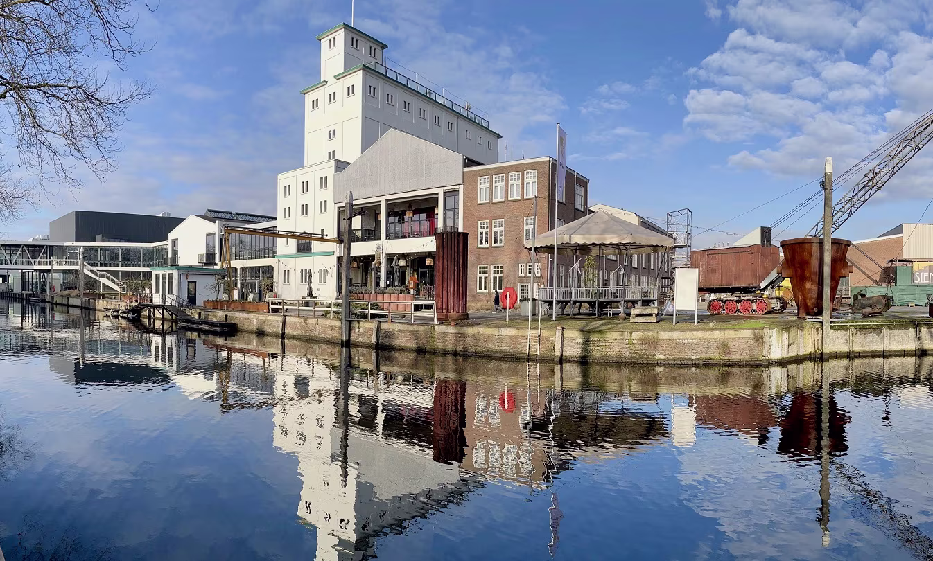 Historische havengebouwen met hoge silo weerspiegelen in het kalme water onder een blauwe hemel met wolken.