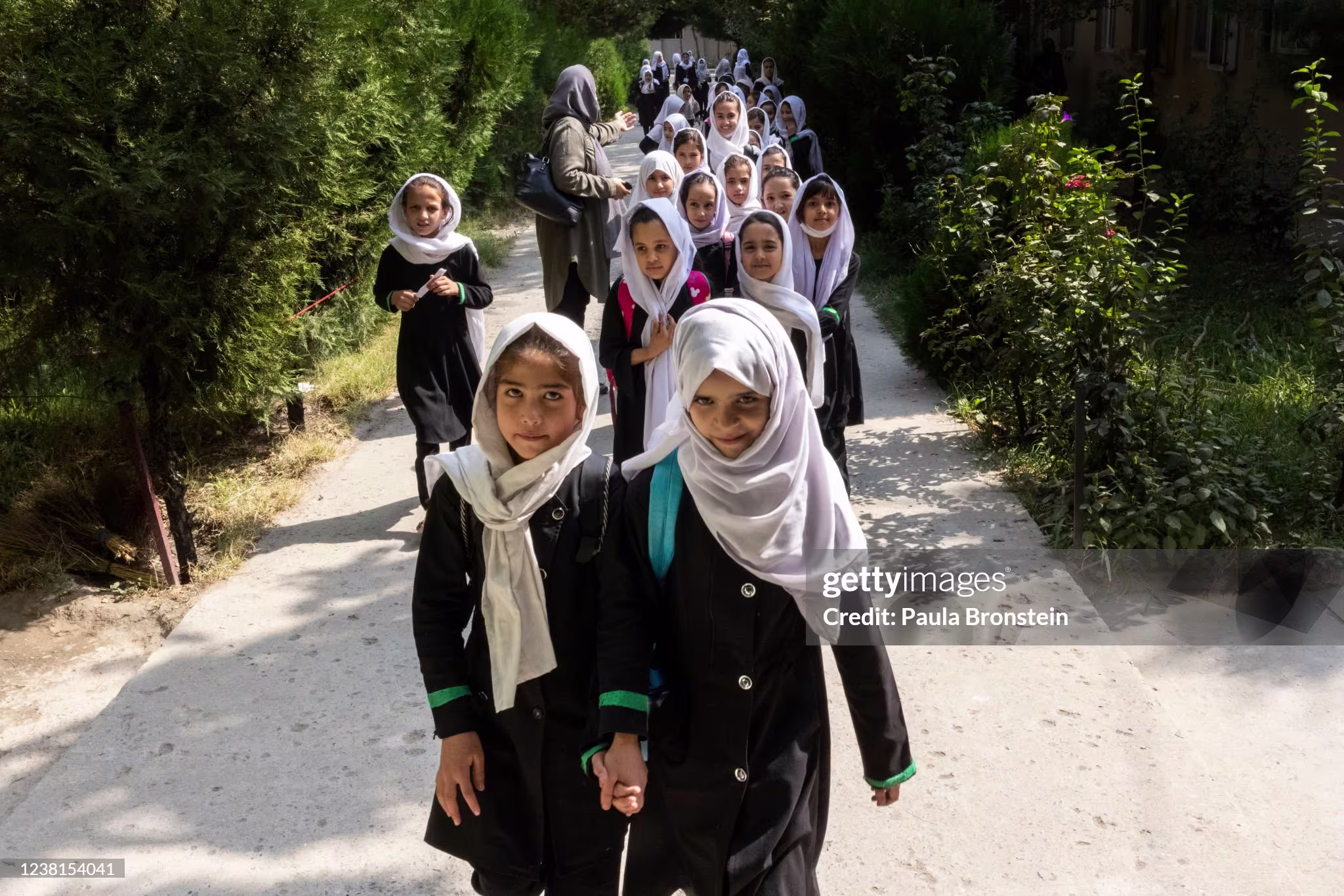 Een groep schoolmeisjes met hoofddoeken loopt in uniform over een pad tussen groen, begeleid door een volwassene.