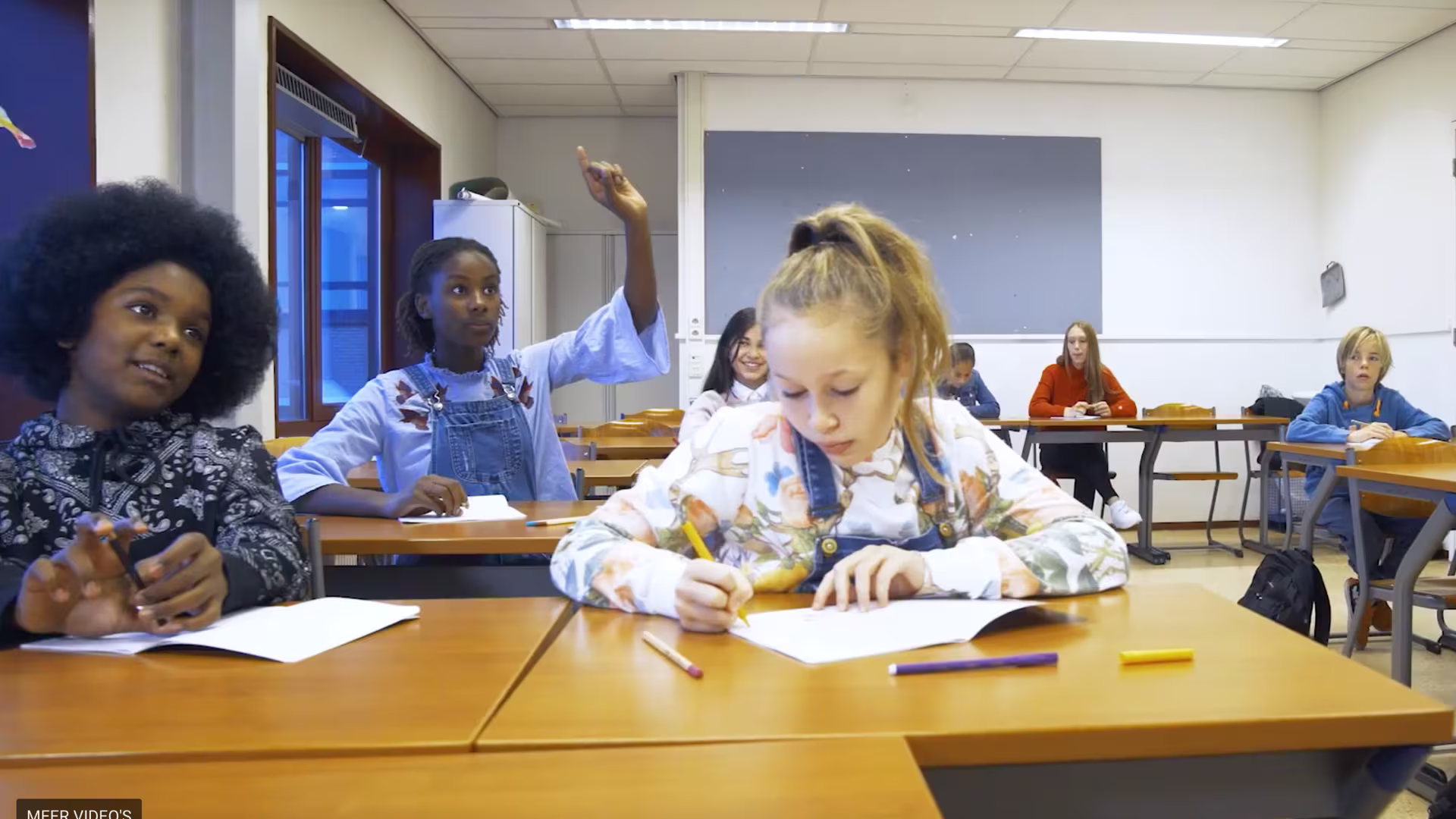 Leerlingen werken in een klaslokaal, één steekt haar hand op terwijl anderen aan hun tafel schrijven en tekenen.