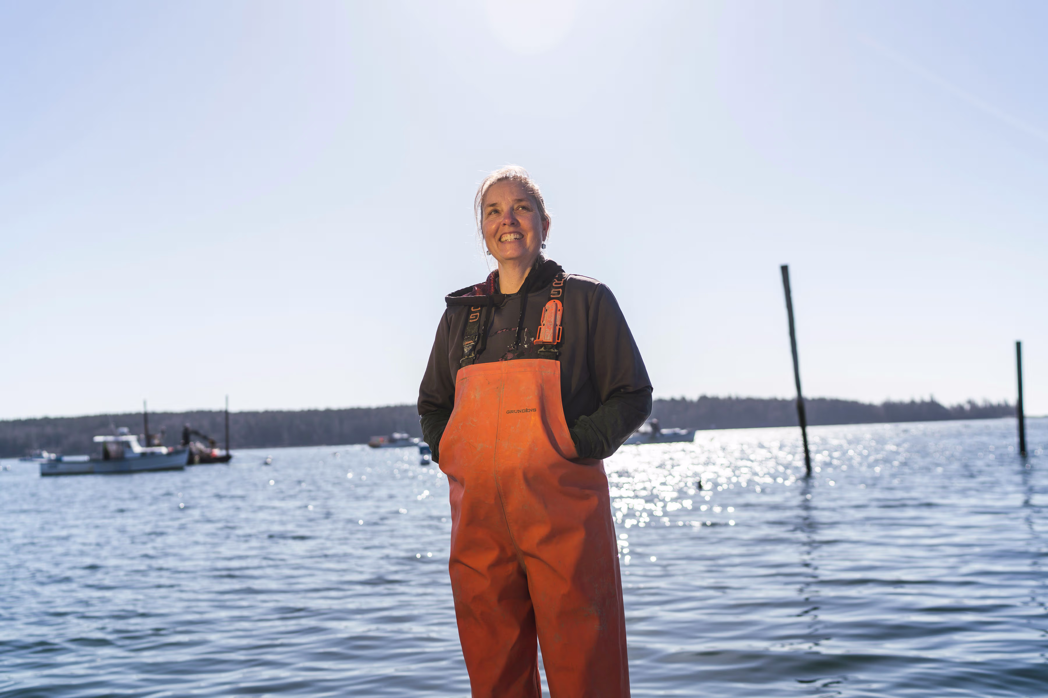Vrouw in oranje werkkleding staat lachend aan de waterkant met boten op de achtergrond.