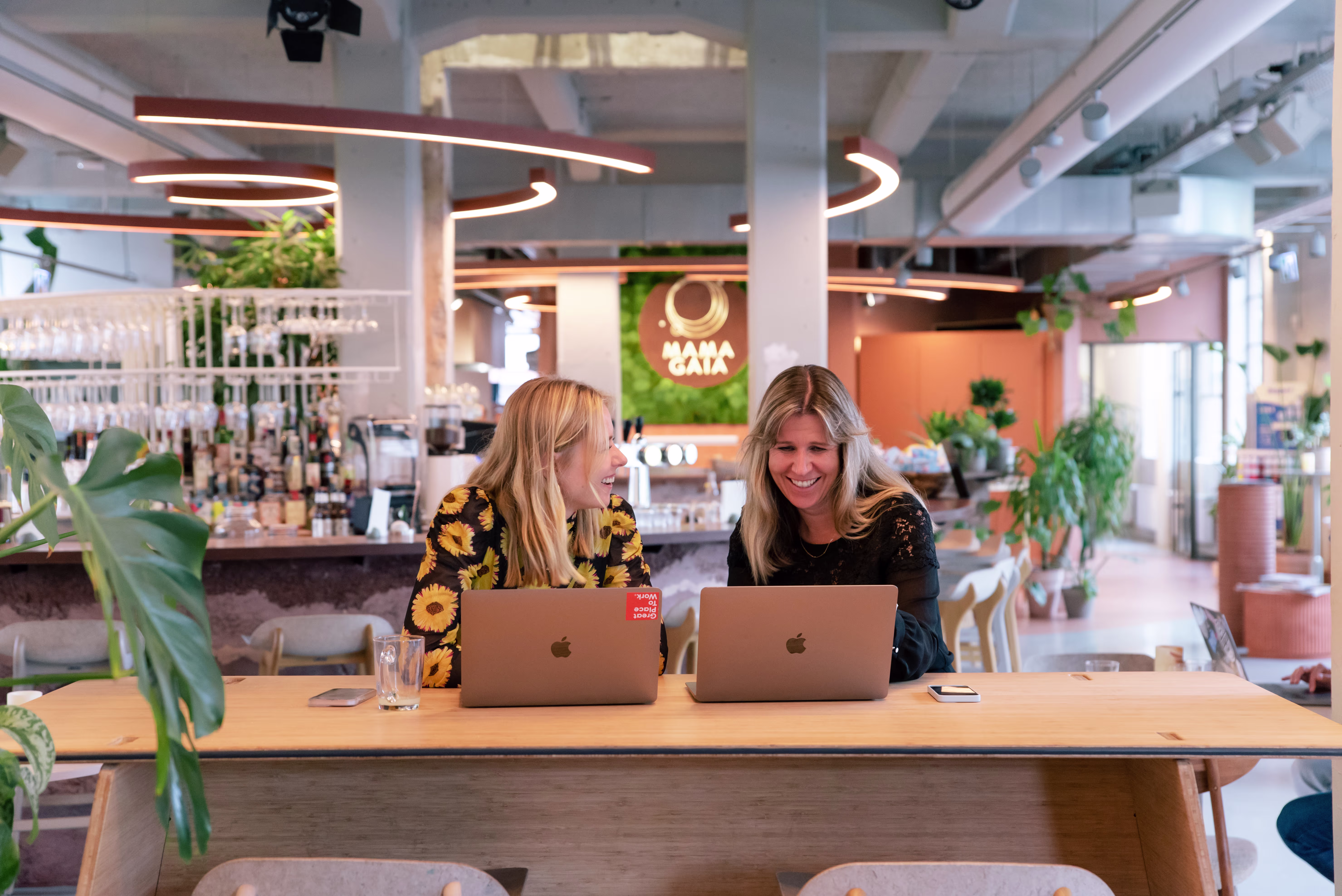 Twee vrouwen werken samen aan laptops in een moderne, groene kantoorruimte met planten en warme verlichting.
