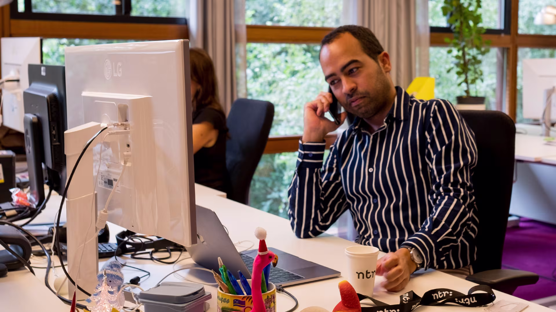Een man in gestreept overhemd belt aan zijn bureau met computer en kantoorspullen, collega op achtergrond.
