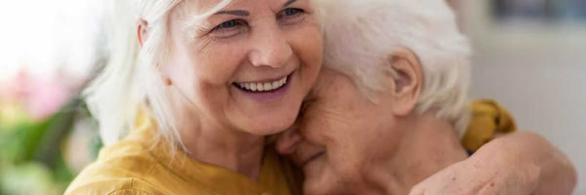 Twee oudere vrouwen met grijs haar omhelzen elkaar liefdevol en glimlachen, wat warmte en zorgzaamheid uitstraalt.