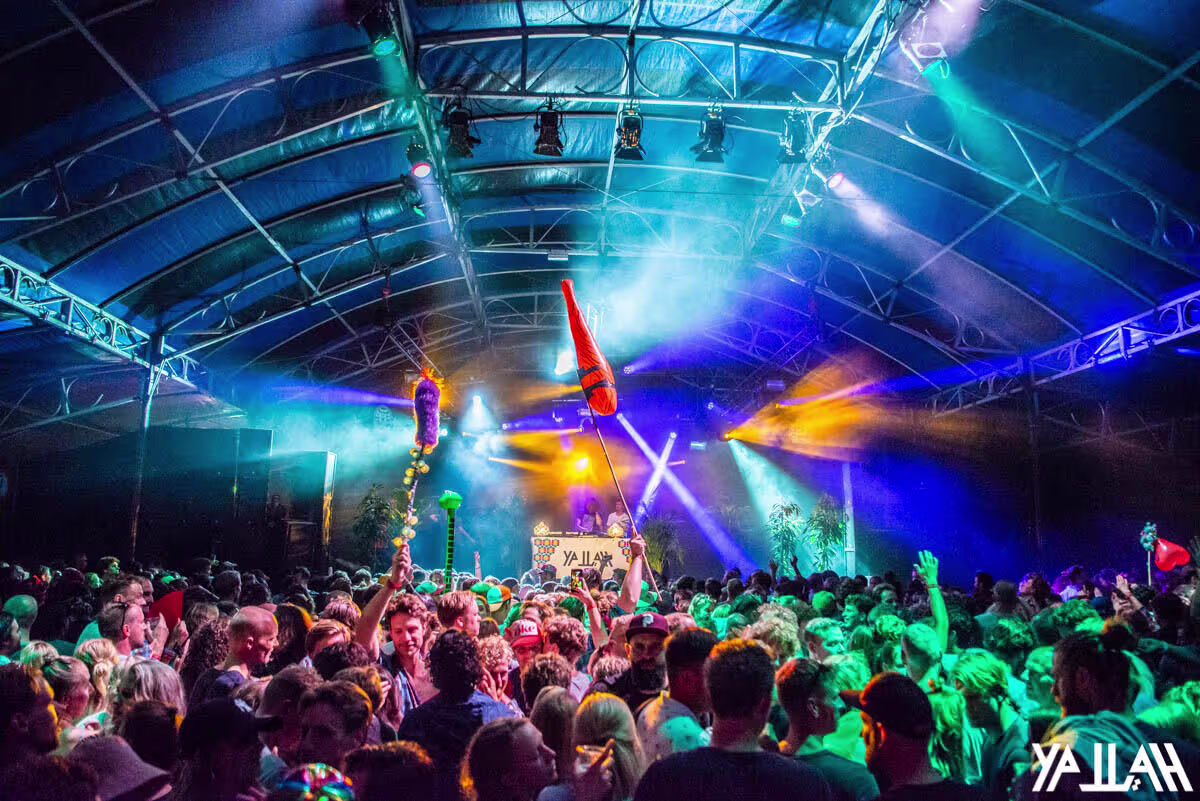 Een vol dansvloer onder kleurrijke lichtshow in een grote evenementenhal met metalen dakconstructie en professionele podiumverlichting.
