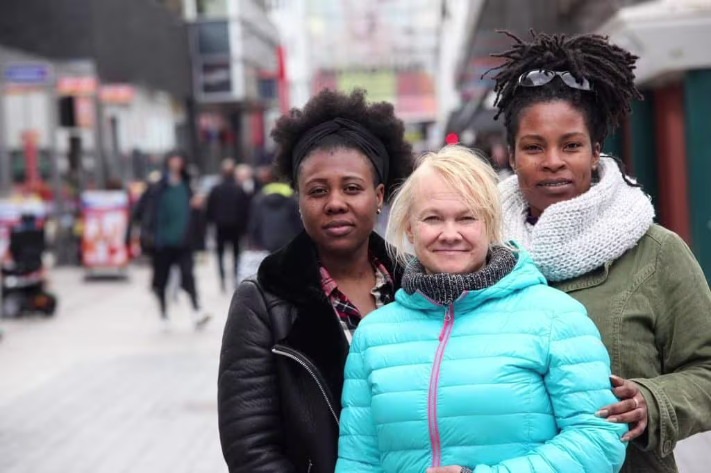 Drie vrouwen staan glimlachend samen op straat, gekleed in winterjassen, met winkels op de achtergrond.