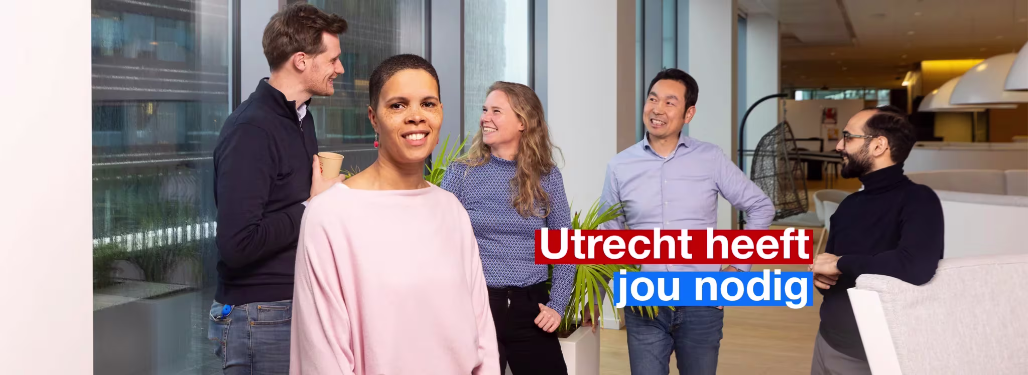 Vijf collega's staan lachend samen in een modern kantoor met grote ramen en hebben een gezellig gesprek.