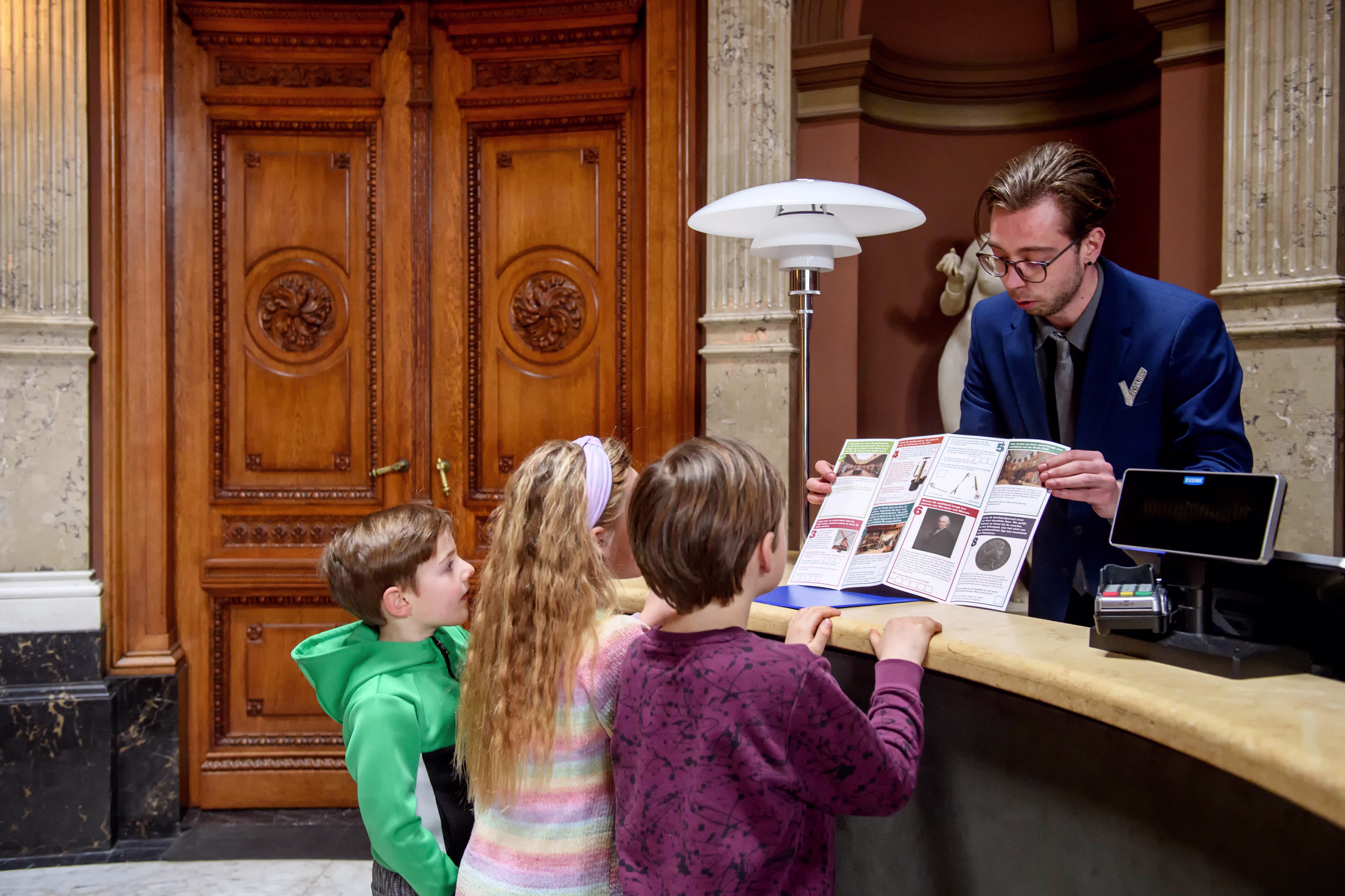 Een medewerker in blauw pak toont drie kinderen een informatiebrochure bij de balie van het museum met klassieke zuilen.