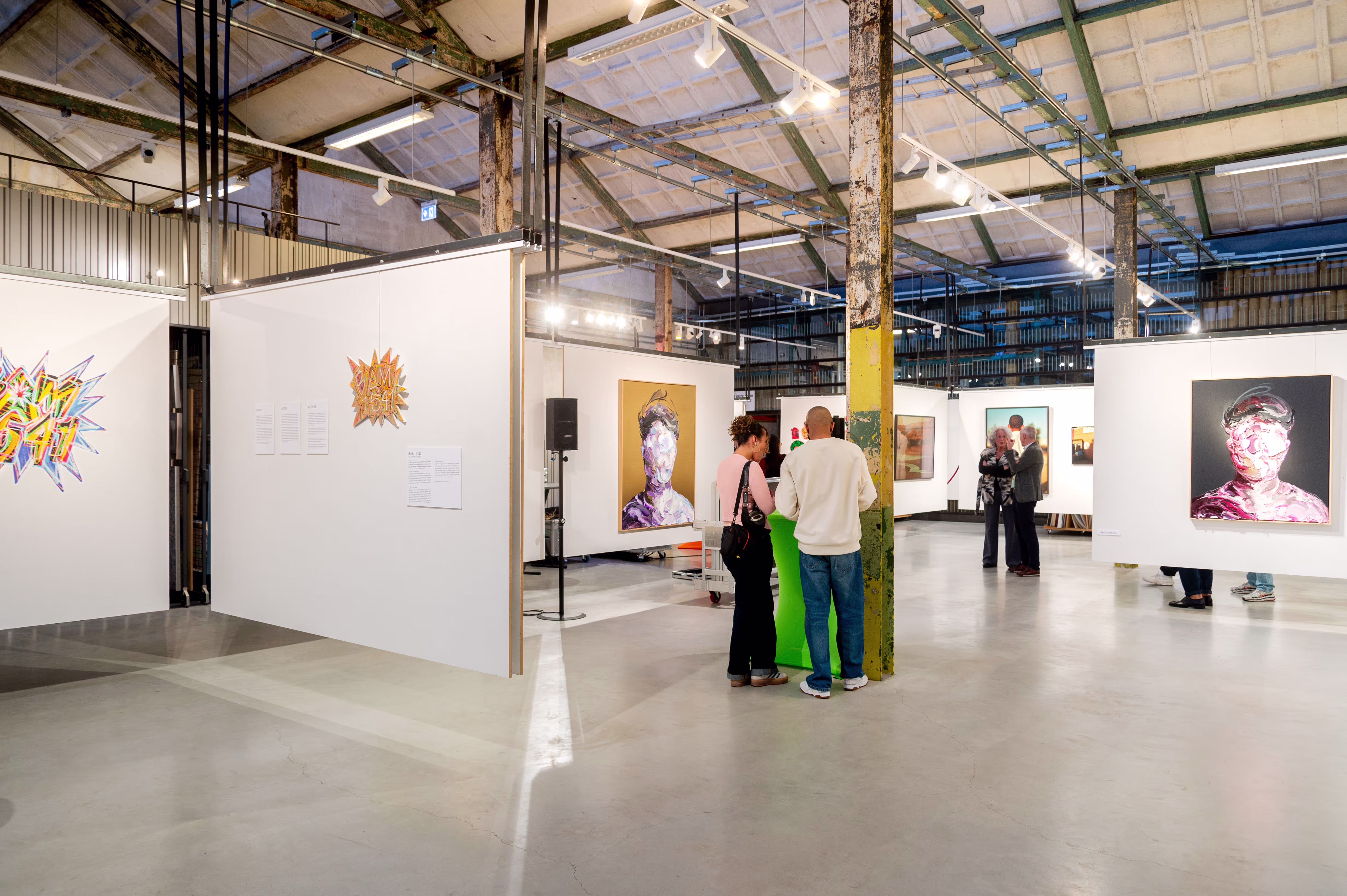 Bezoekers bekijken kunstwerken in een moderne galerie met industriële architectuur en witte tentoonstellingswanden.