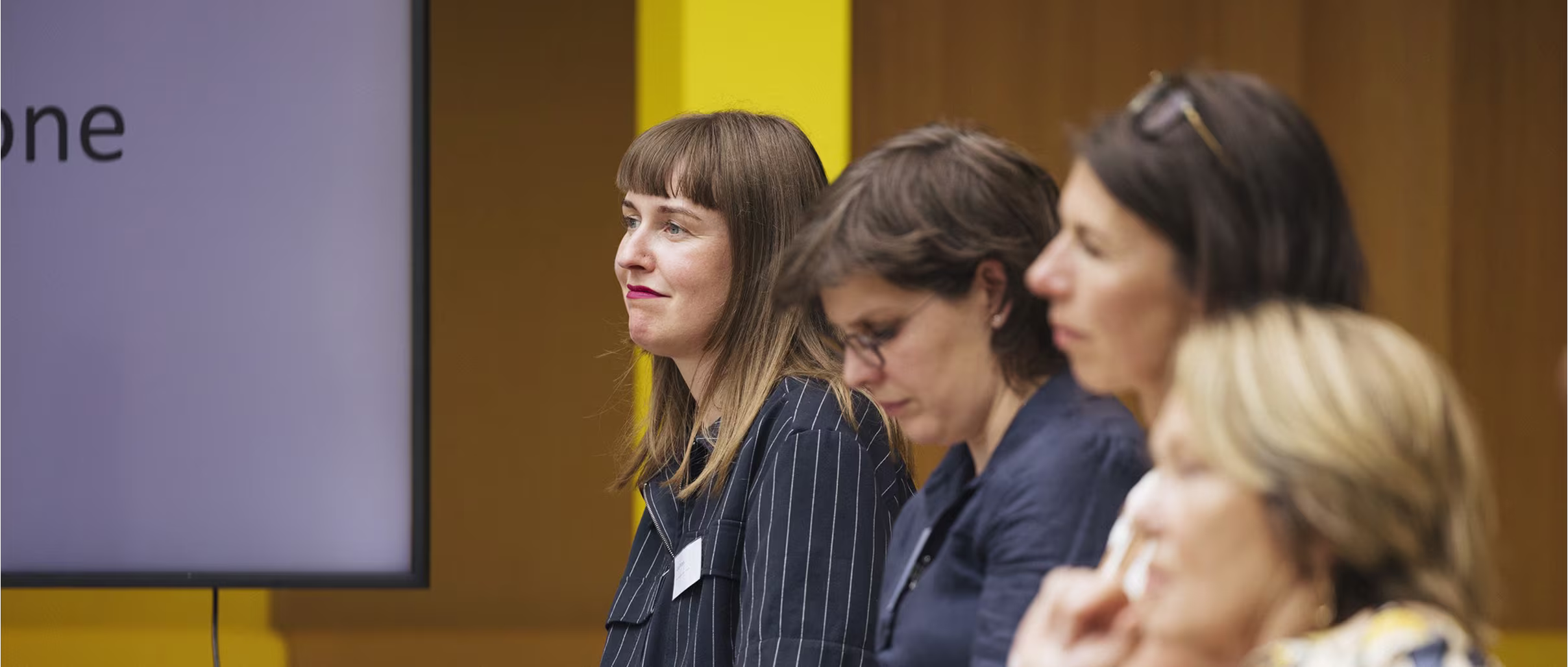 Groep professionele vrouwen in zakelijke kleding tijdens een presentatie of bijeenkomst, met scherm op de achtergrond.