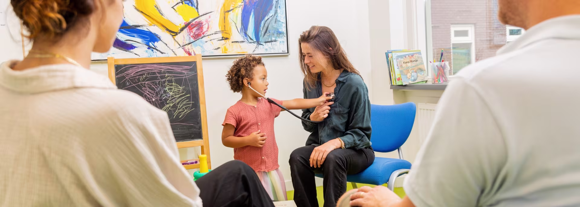 Een vrouw in gesprek met een kind in een kleurrijke ruimte, terwijl twee volwassenen toekijken.