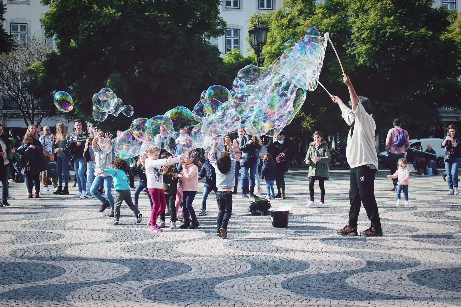 Een straatartiest maakt grote zeepbellen op een stenen plein terwijl kinderen en volwassenen toekijken en meespelen.