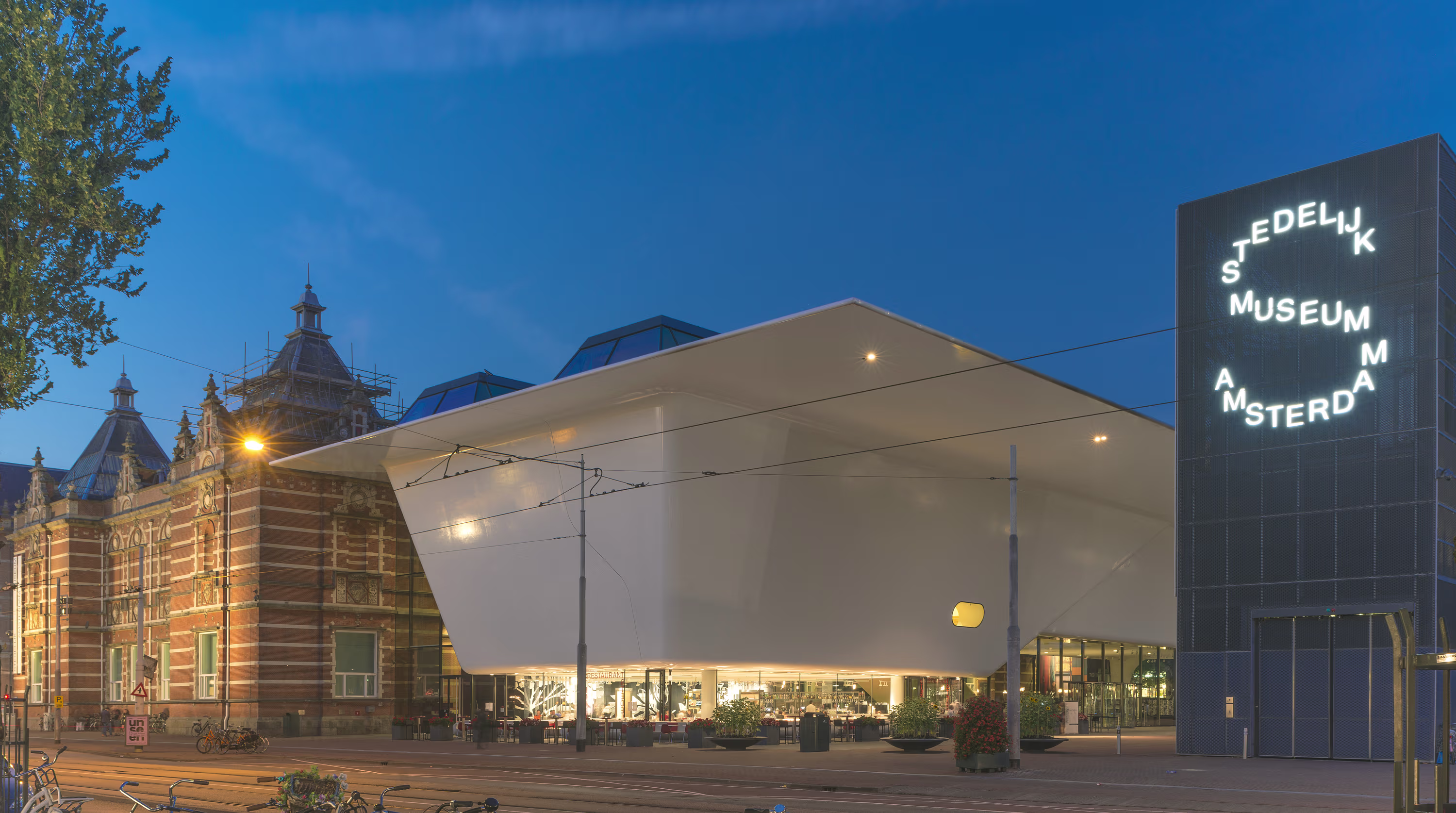 Avondfoto van het Stedelijk Museum Amsterdam met zijn moderne witte aanbouw naast het historische gebouw, verlicht tegen een blauwe hemel.