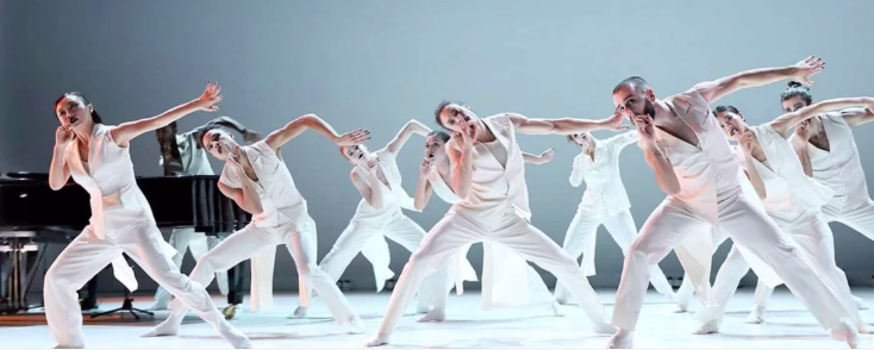 Een groep dansers in witte kleding voert een gecoördineerde moderne dans uit op een podium met een piano op de achtergrond.