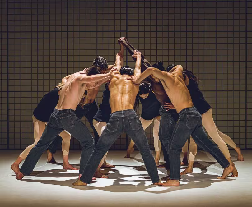Groep shirtloze mannelijke dansers in spijkerbroeken vormt cirkel met armen omhoog in theatrale dansvoorstelling.
