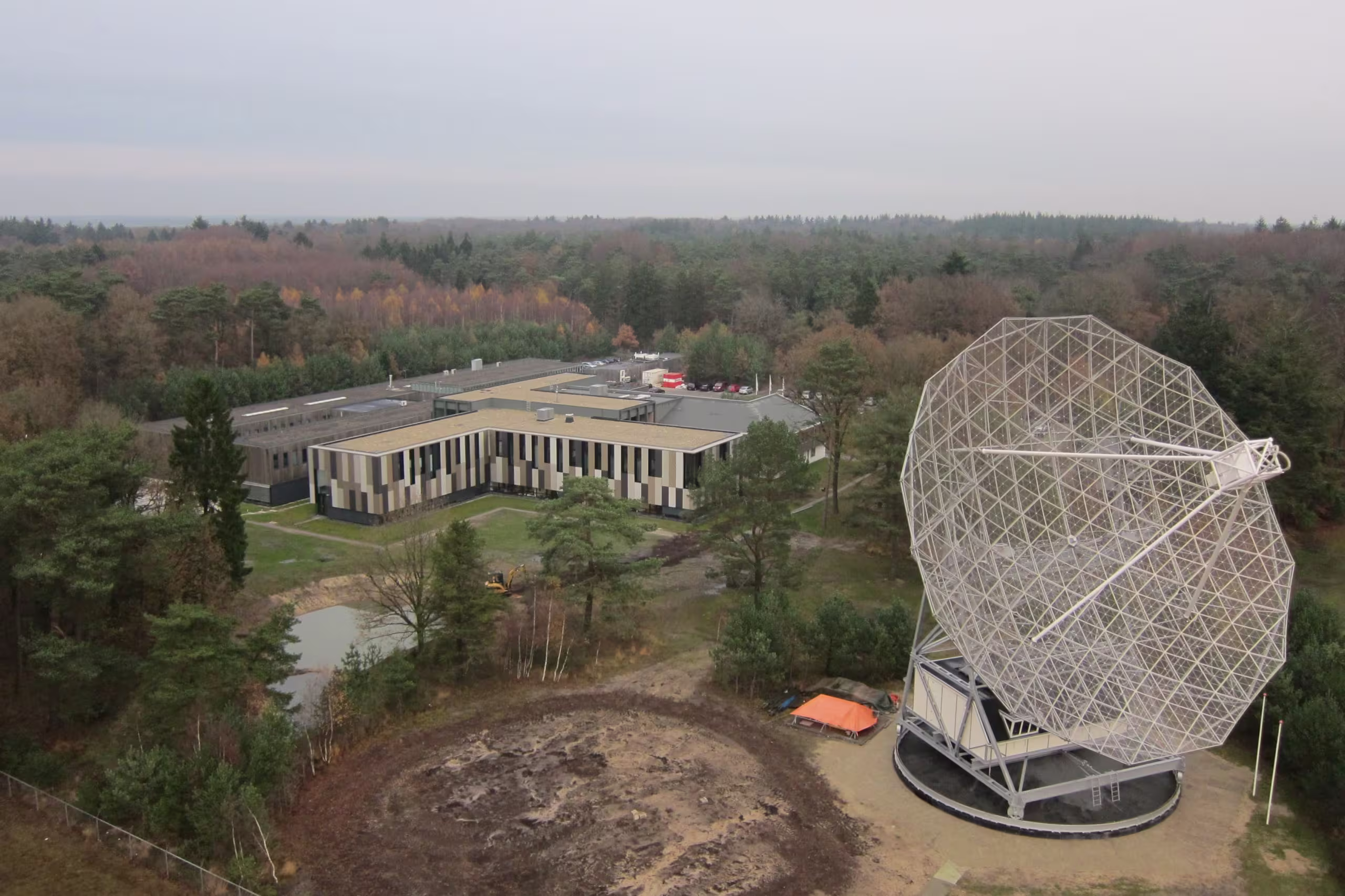 Luchtfoto van ASTRON-complex met moderne gebouwen en grote witte radiotelescoop omringd door bossen.