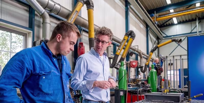Twee mannen in werkkleding bespreken technische apparatuur in een industriële werkplaats met groene machines en leidingen.