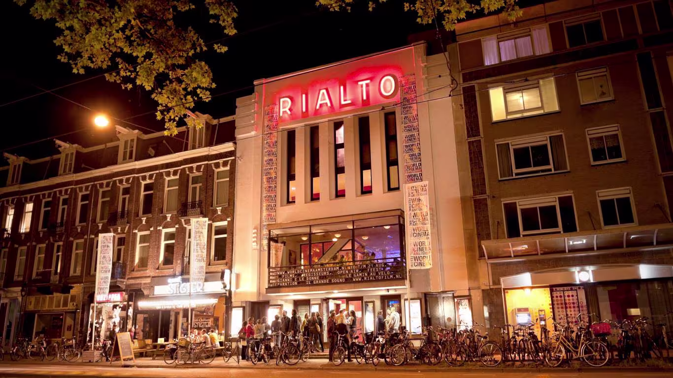 Nachtfoto van filmhuis Rialto in Amsterdam met verlichte gevel, mensen en fietsen voor de ingang.