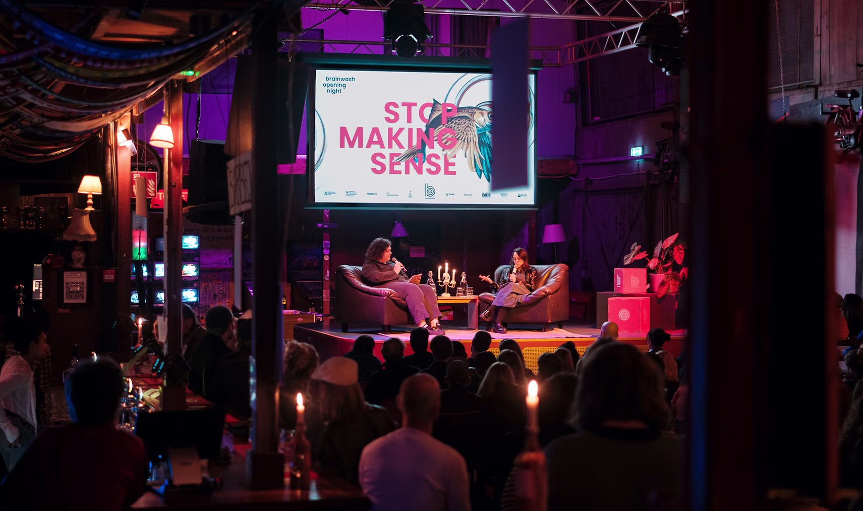 Een evenement in een sfeervolle zaal met publiek dat luistert naar sprekers op een podium onder paarse verlichting.