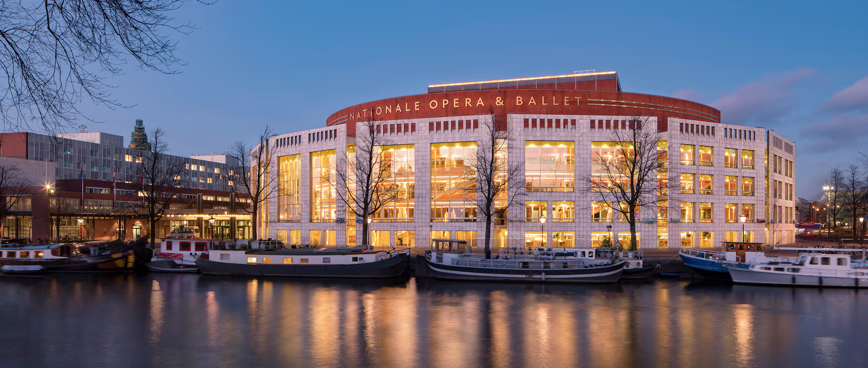 Het verlichte gebouw van De Nationale Opera & Ballet aan het water tijdens de avondschemering, met boten op de voorgrond.