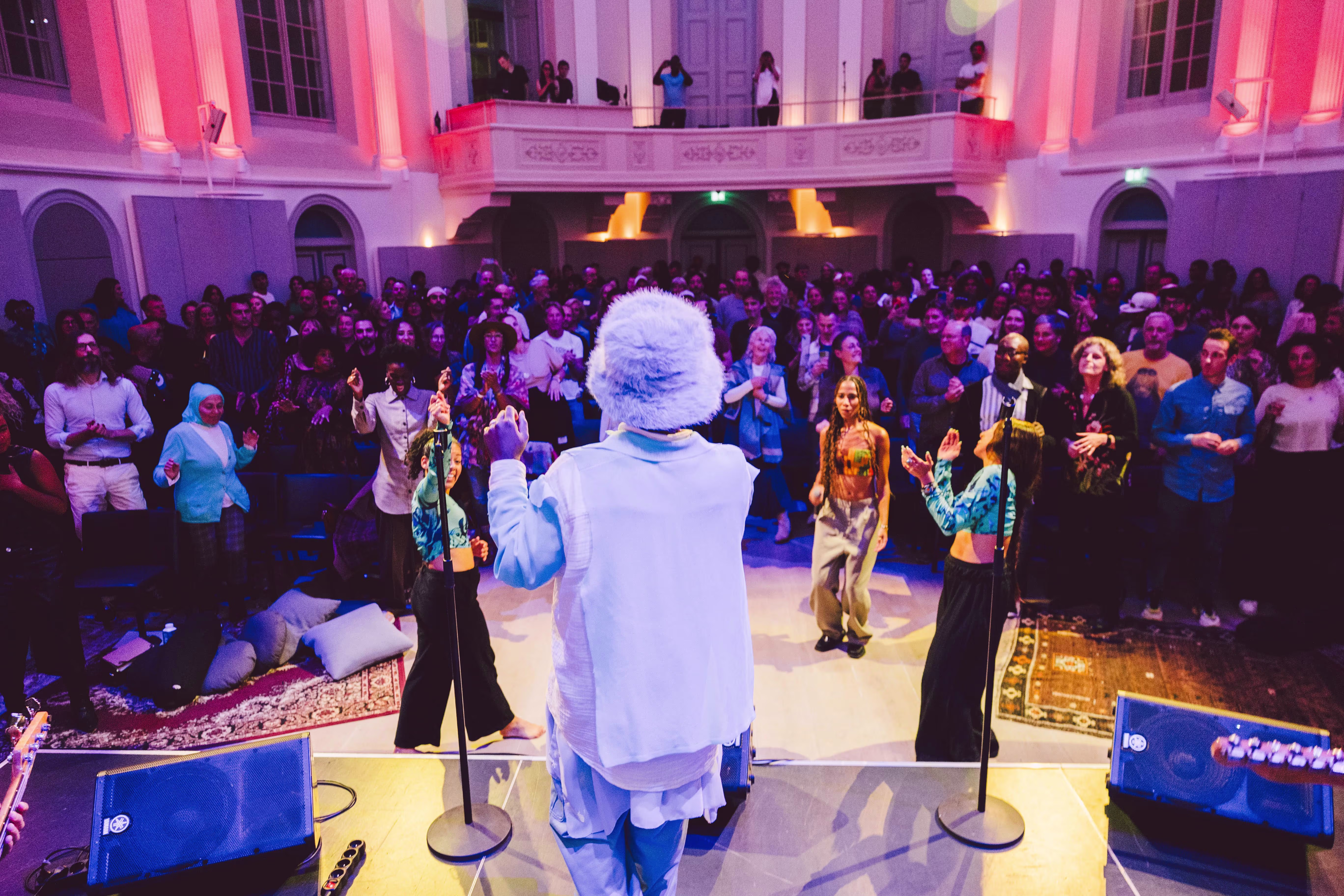 Een performer in wit gewaad staat op een podium voor een grote menigte in een historisch theater met paarse en roze verlichting.