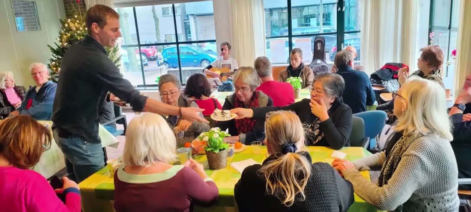 Een man serveert eten aan ouderen die aan tafels zitten in een gezellige ruimte met kerstboom op de achtergrond.
