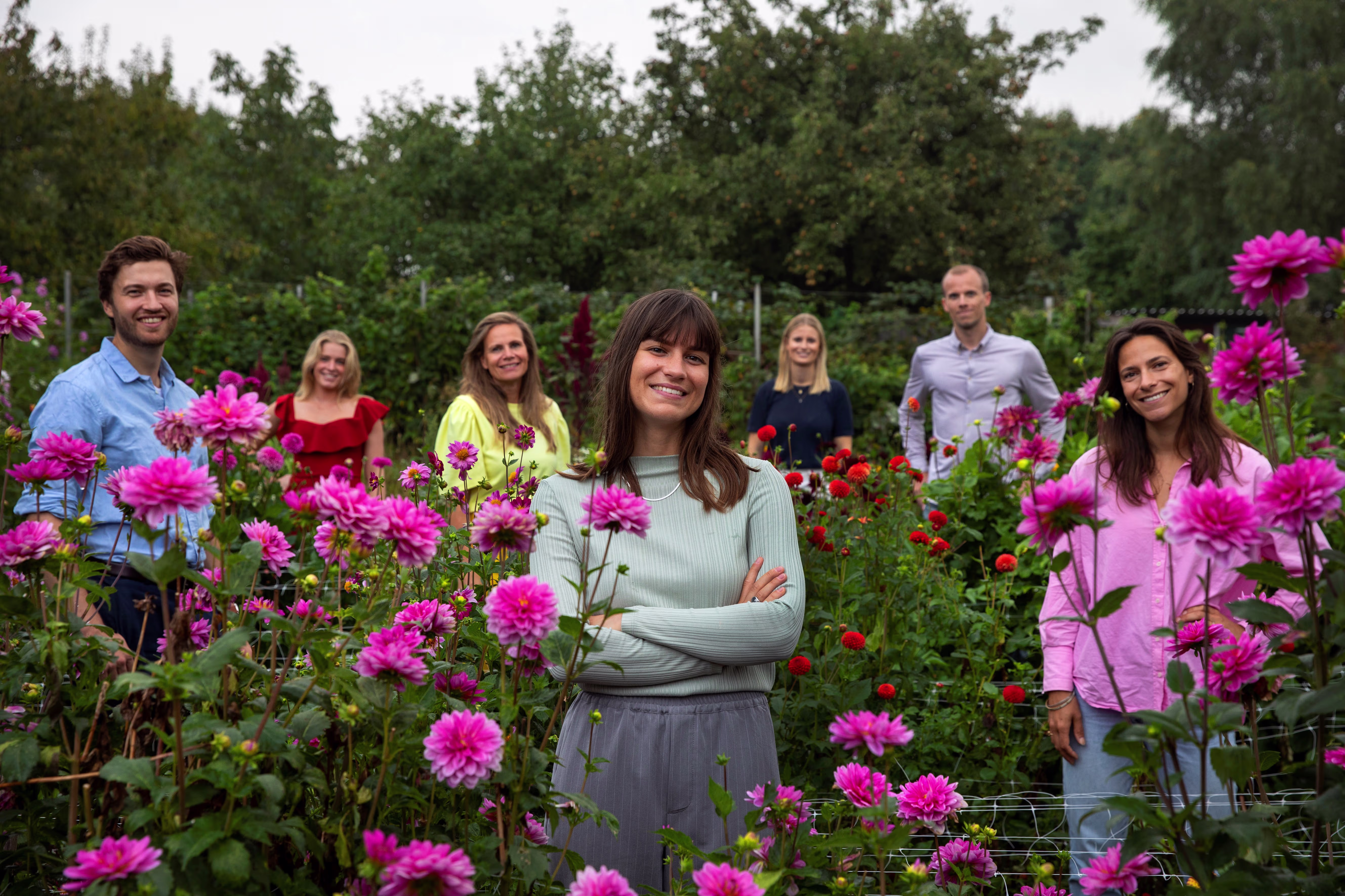 Een groep van zeven collega's staat lachend tussen kleurrijke bloemen in een tuin, met de focus op een vrouw vooraan.