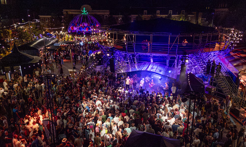 Groot openlucht theaterfestival met honderden bezoekers rond een verlichte podiumopstelling met blauwe belichting 's avonds.