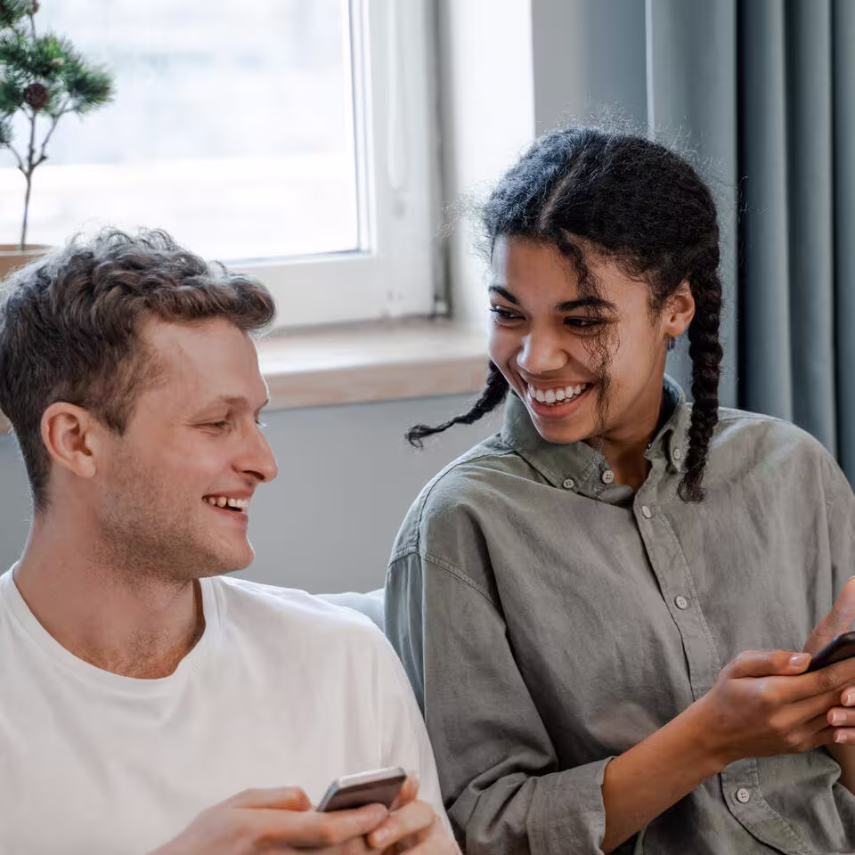 Een lachende man en vrouw zitten naast elkaar met smartphones in hun handen en kijken elkaar vrolijk aan bij een raam.