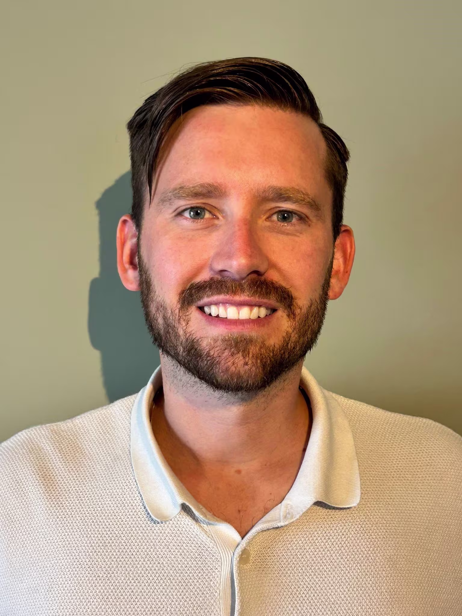 A smiling man with a beard, wearing a white shirt, poses for a professional portrait against a neutral background.