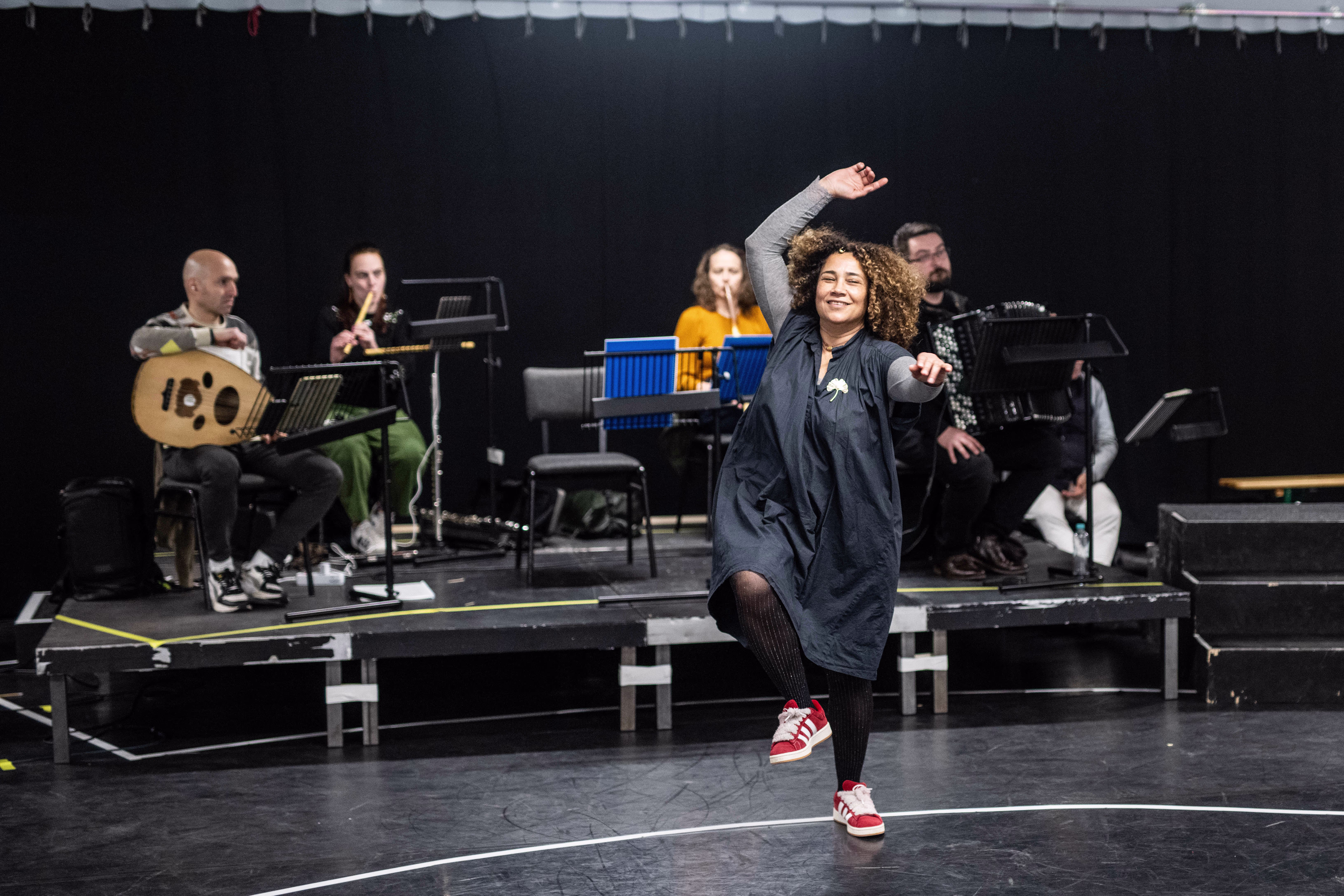 Een danseres in een grijze jurk danst voor een orkest op een zwart podium tijdens een repetitie of voorstelling.