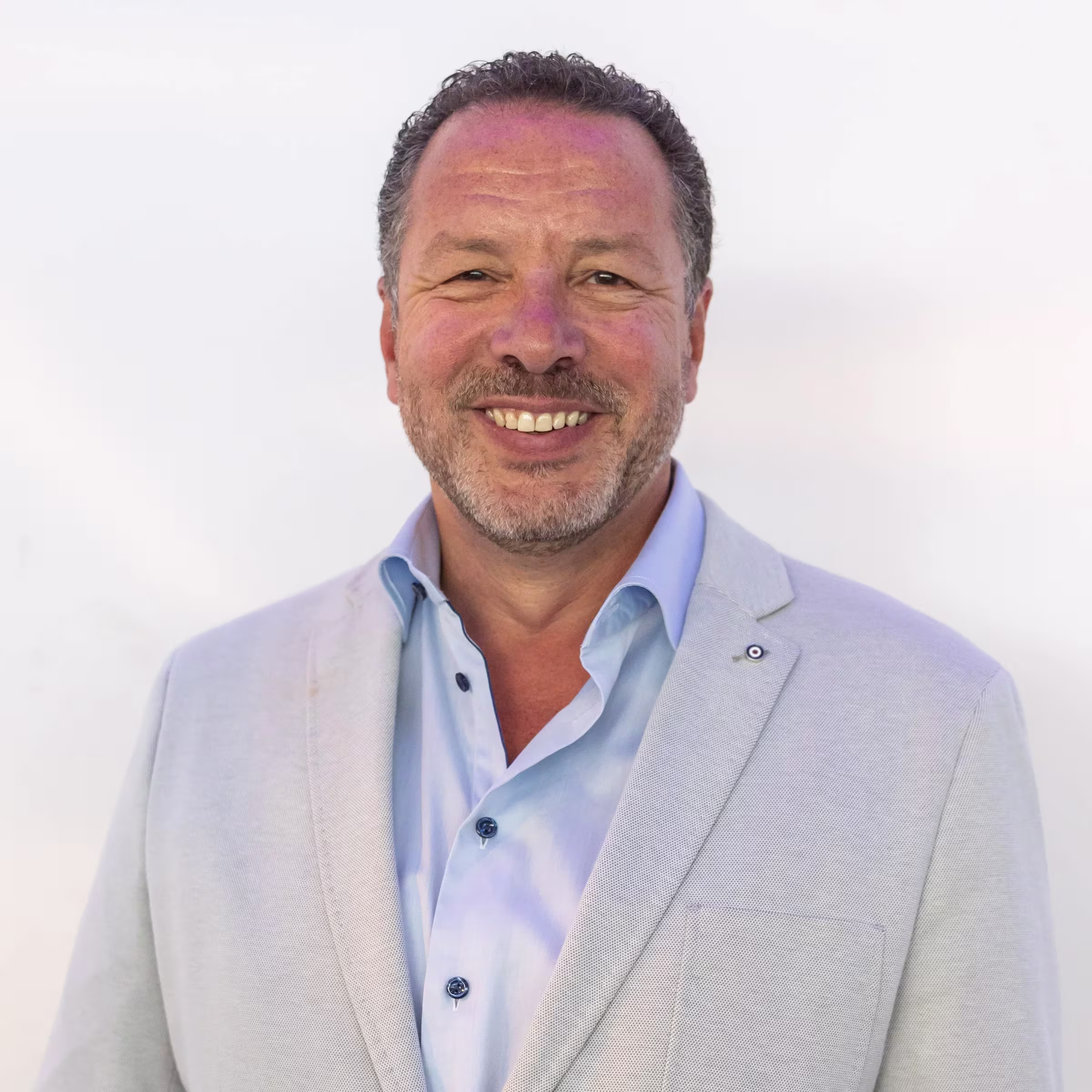 A smiling man in a light gray jacket and light blue shirt against a white background, a professional portrait photo.