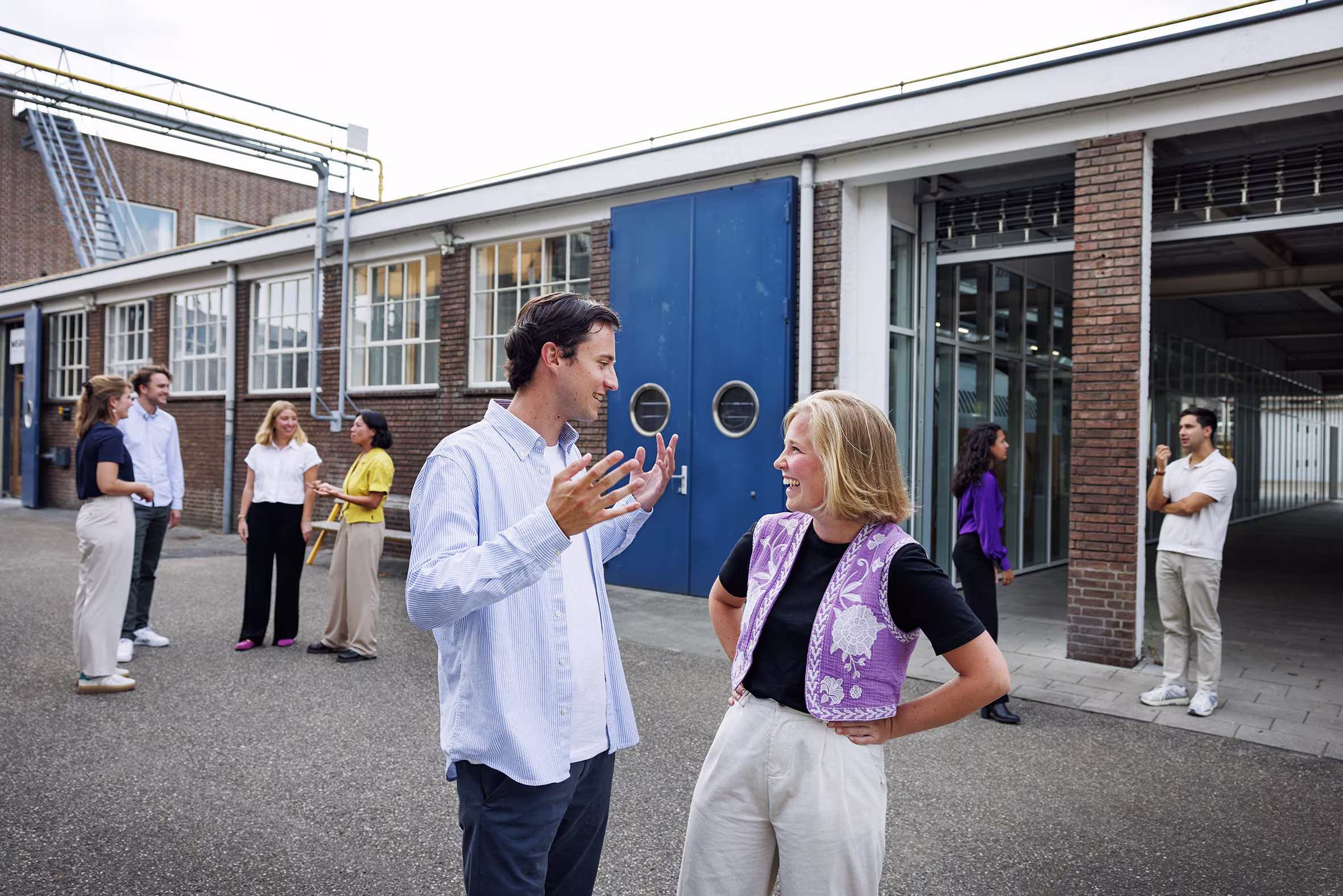 Een man in blauw overhemd praat enthousiast met een blonde vrouw in paars vest, op de achtergrond staan meer collega's.
