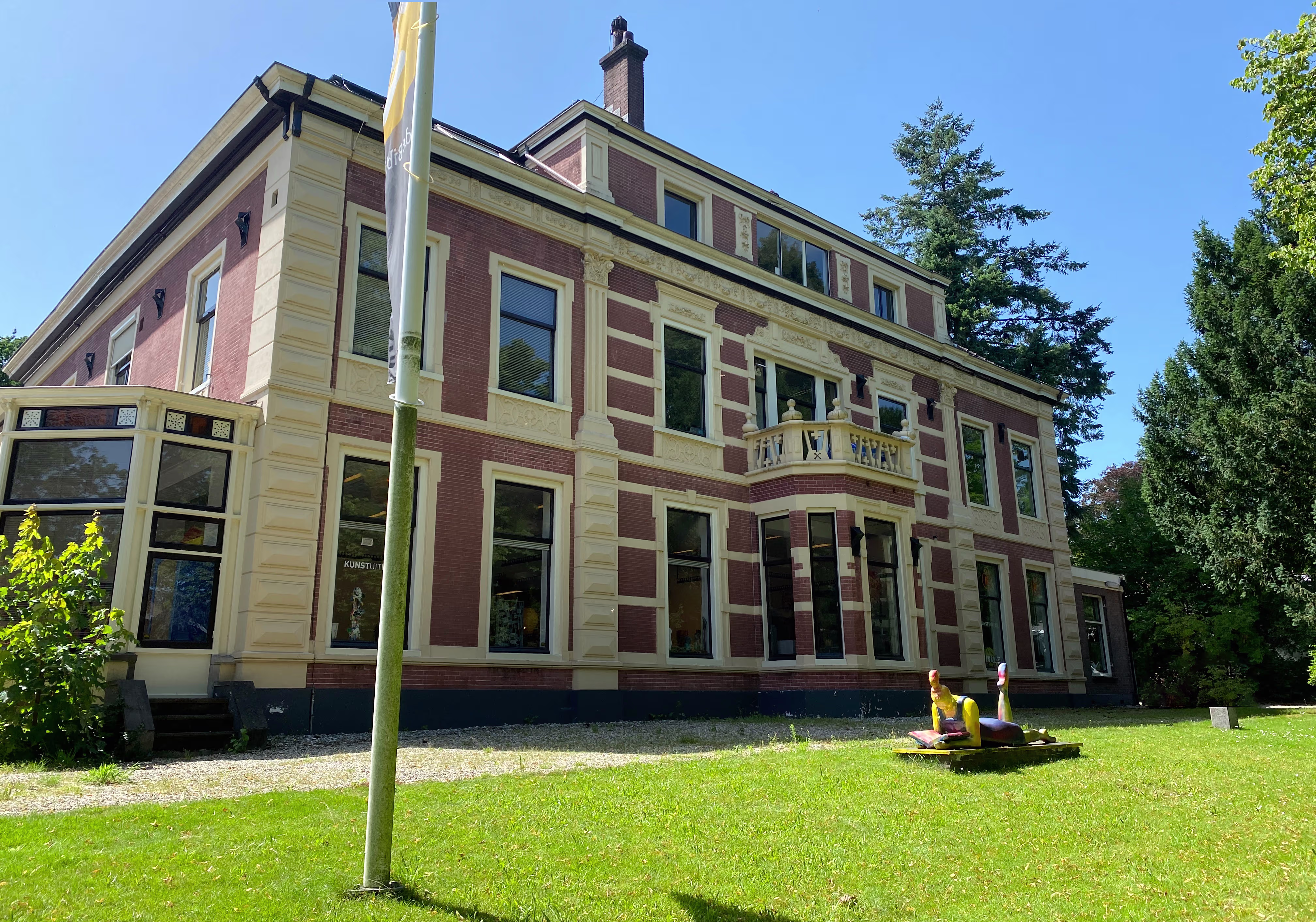 Historisch bakstenen gebouw met lichtgele accenten, omgeven door groen gras en bomen onder blauwe hemel.