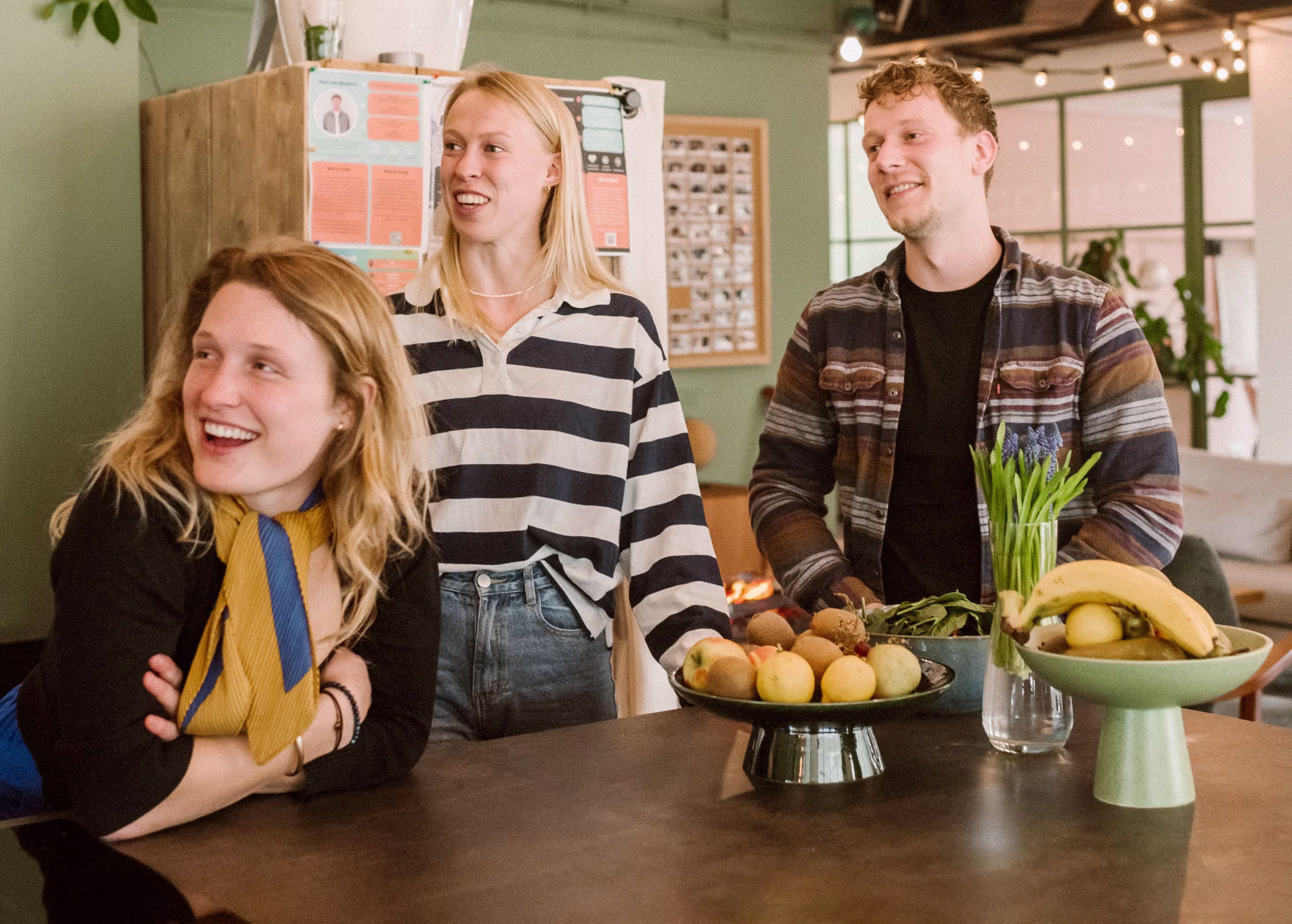 Drie lachende collega's staan rond een tafel met fruit in een moderne kantooromgeving met groene muren en string lights.