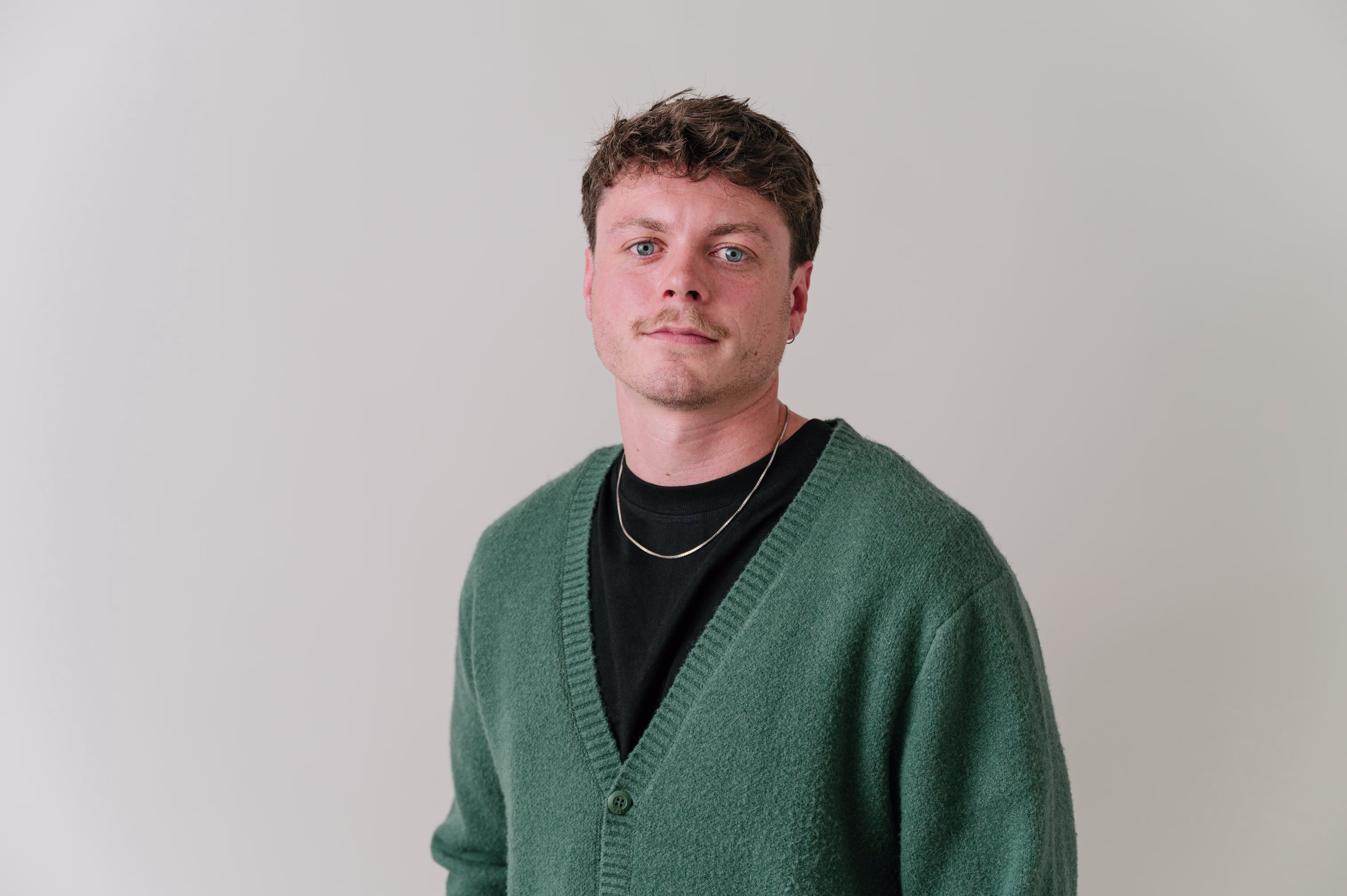 Een jonge man met krullend haar draagt een groene cardigan over een zwart shirt en kijkt vriendelijk in de camera.
