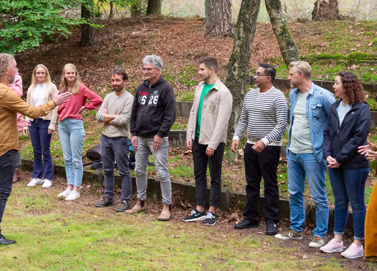 Groep collega's staat in een rij in een bosrijke omgeving, glimlachend tijdens een teamuitje of bedrijfsactiviteit.