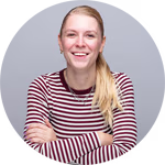 A smiling woman with blonde hair wearing a red-and-white striped sweater, sitting with her arms crossed against a gray background.