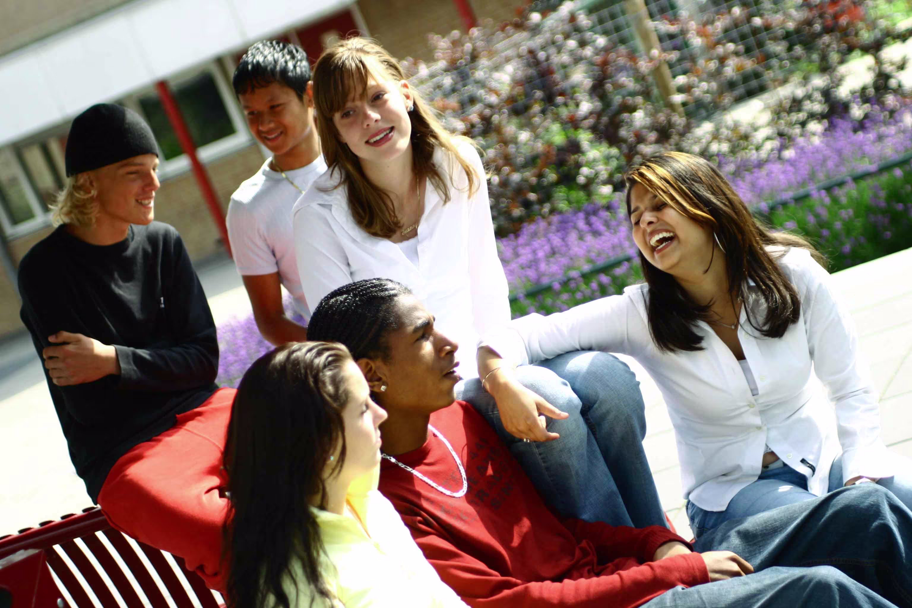 Groep jonge mensen zit lachend samen op rode banken buiten op een schoolplein of universiteitslocatie.