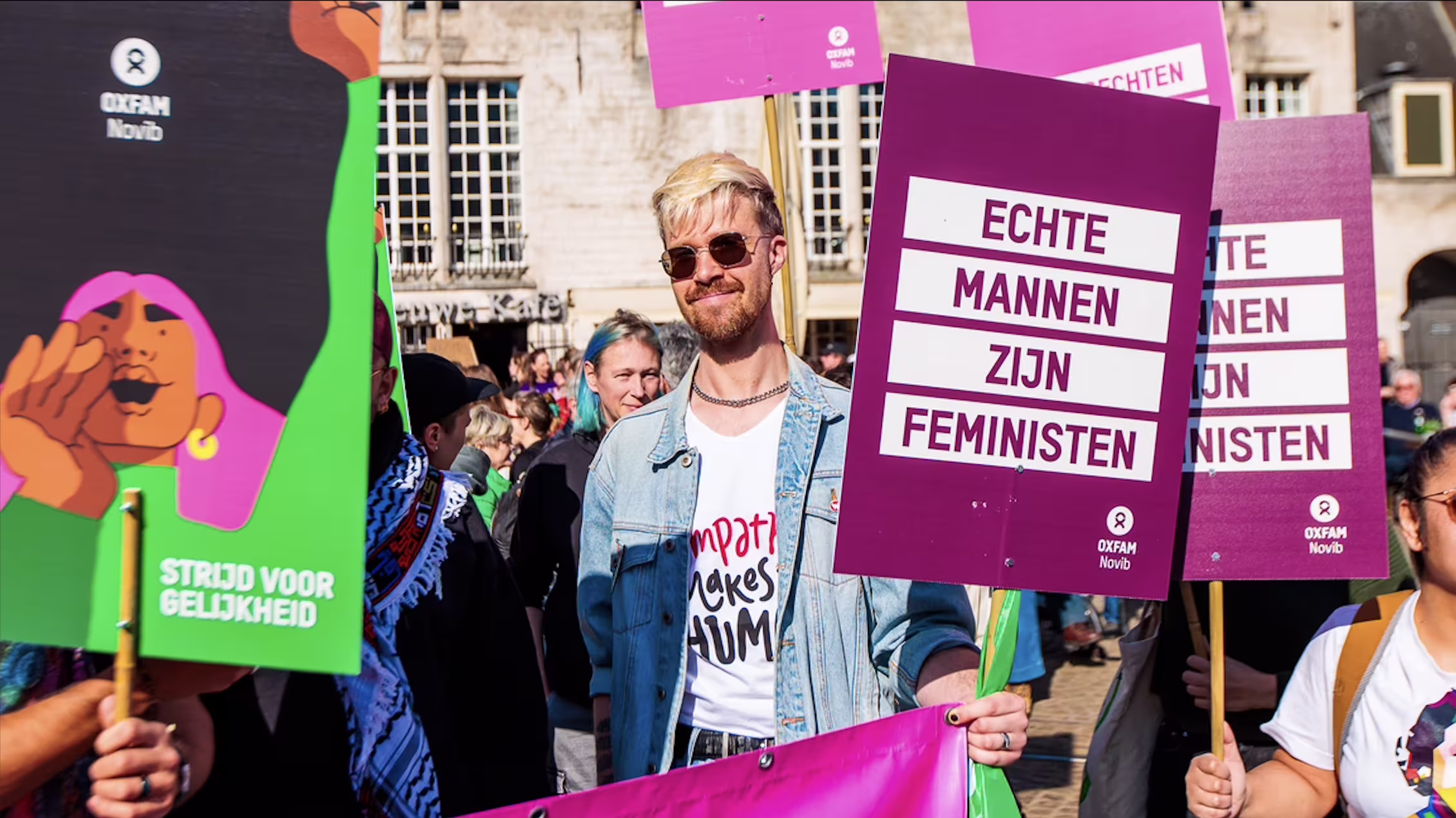 Man met zonnebril en denim jasje houdt paars protest bord vast met tekst "ECHTE MANNEN ZIJN FEMINISTEN" tijdens demonstratie.