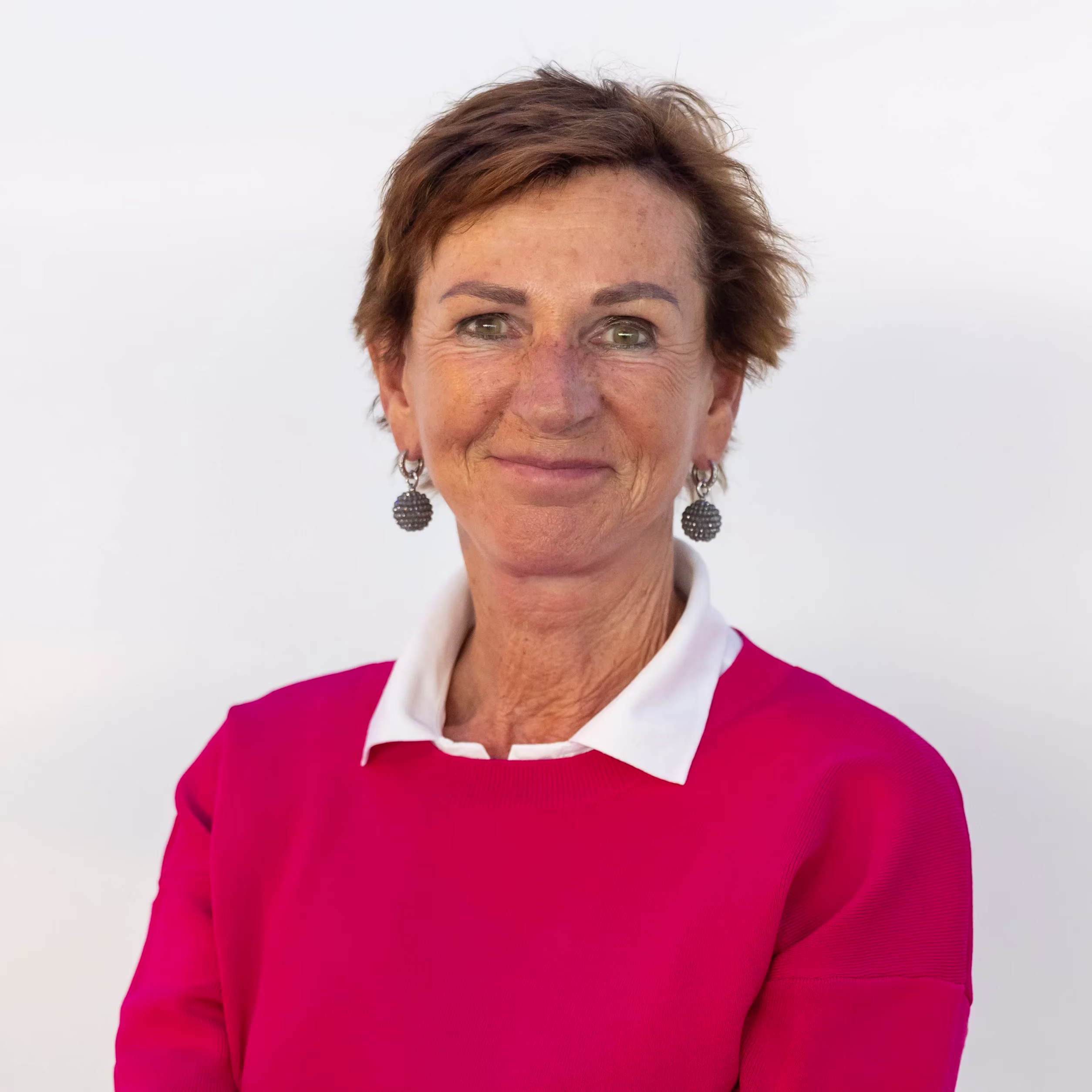 A professional woman with short brown hair is wearing a fuchsia-pink sweater with a white collar and hoop earrings.