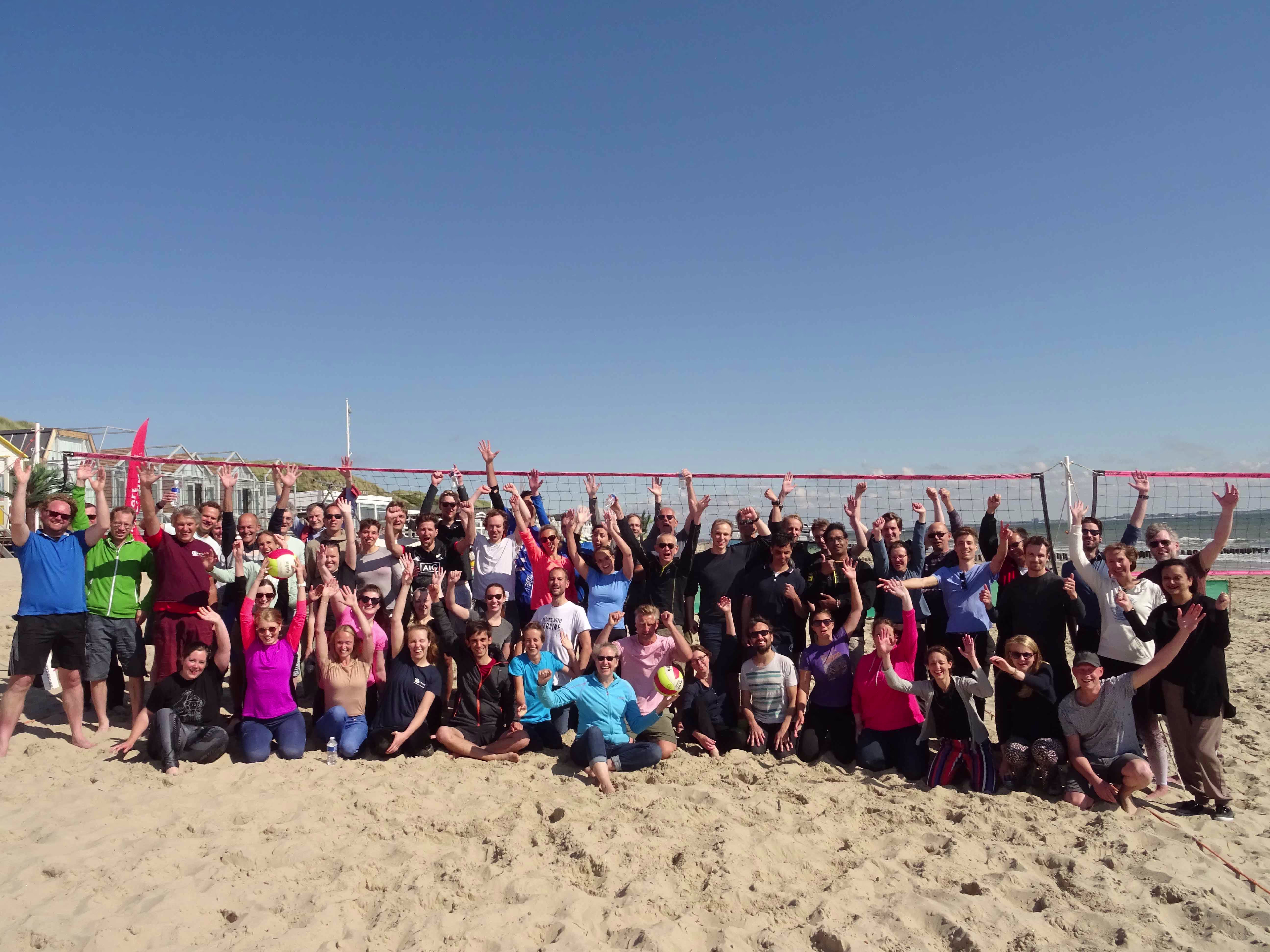 Grote groep collega's poseert enthousiast met opgestoken armen op het strand voor een beachvolleybalnet.