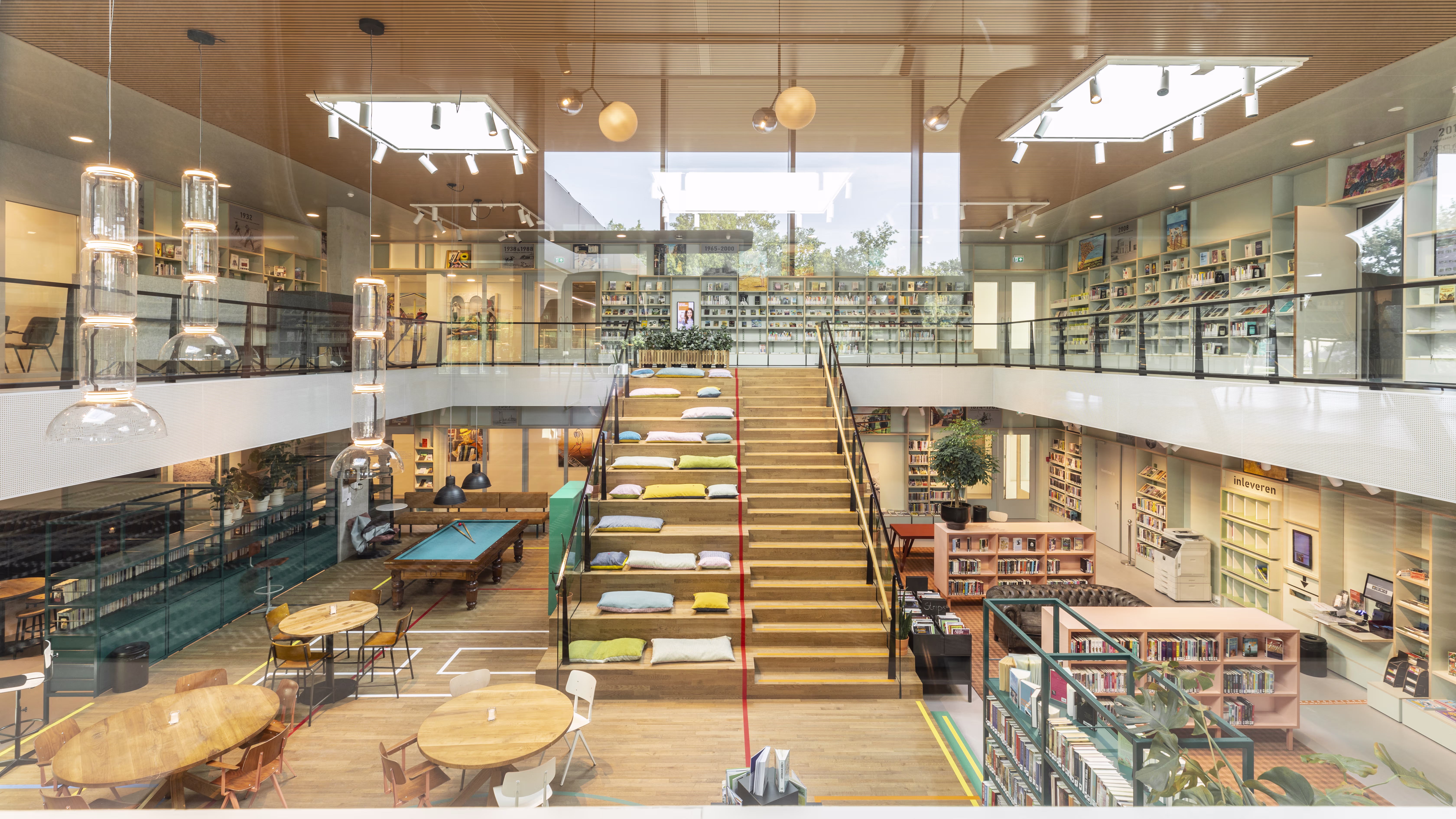 A modern library with a glass staircase, bookshelves on two floors, seating areas, and natural light streaming in through large windows.